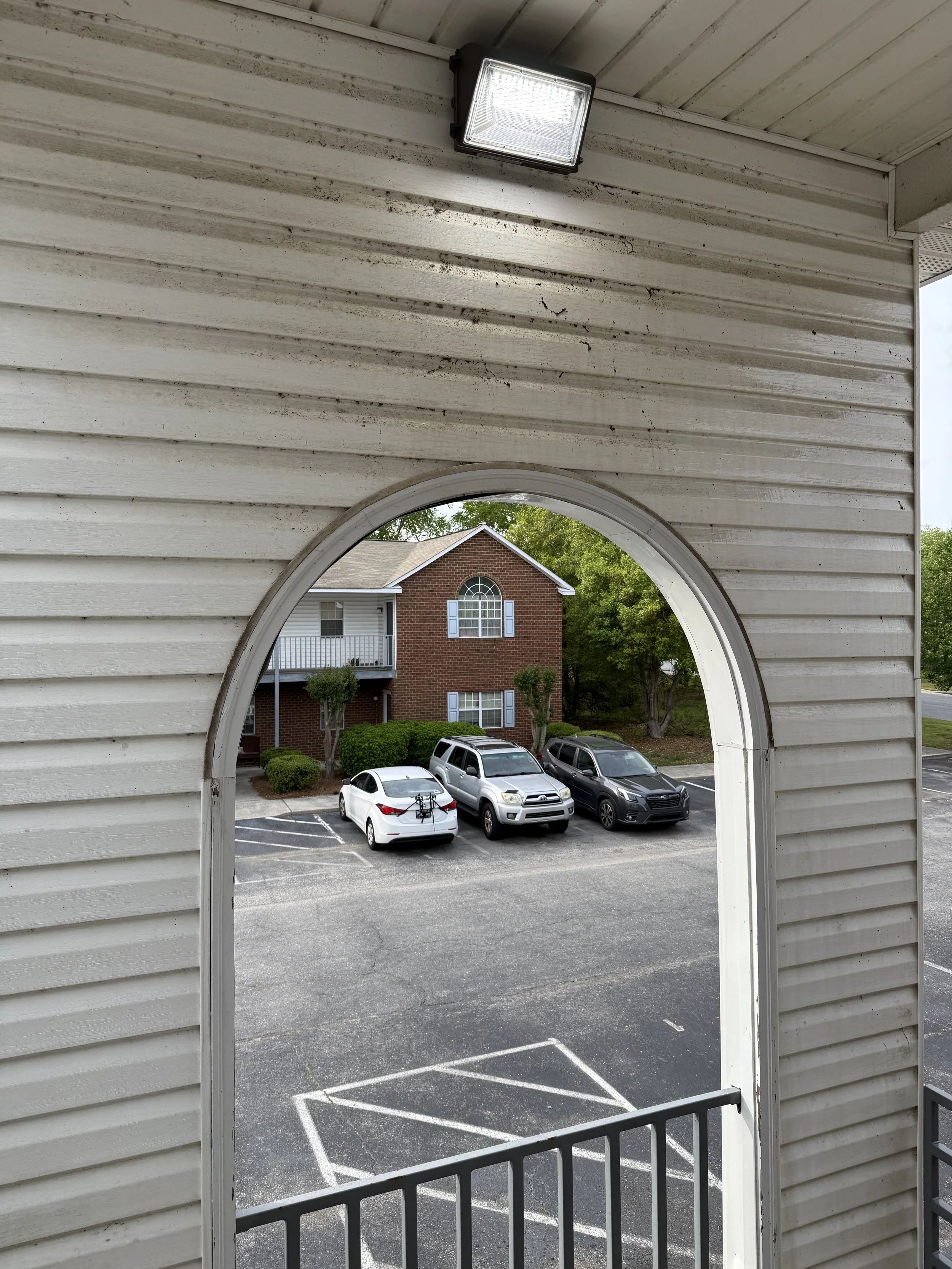 View of a parking lot through an arched window in a white wooden building exterior with a light fixture on the ceiling.