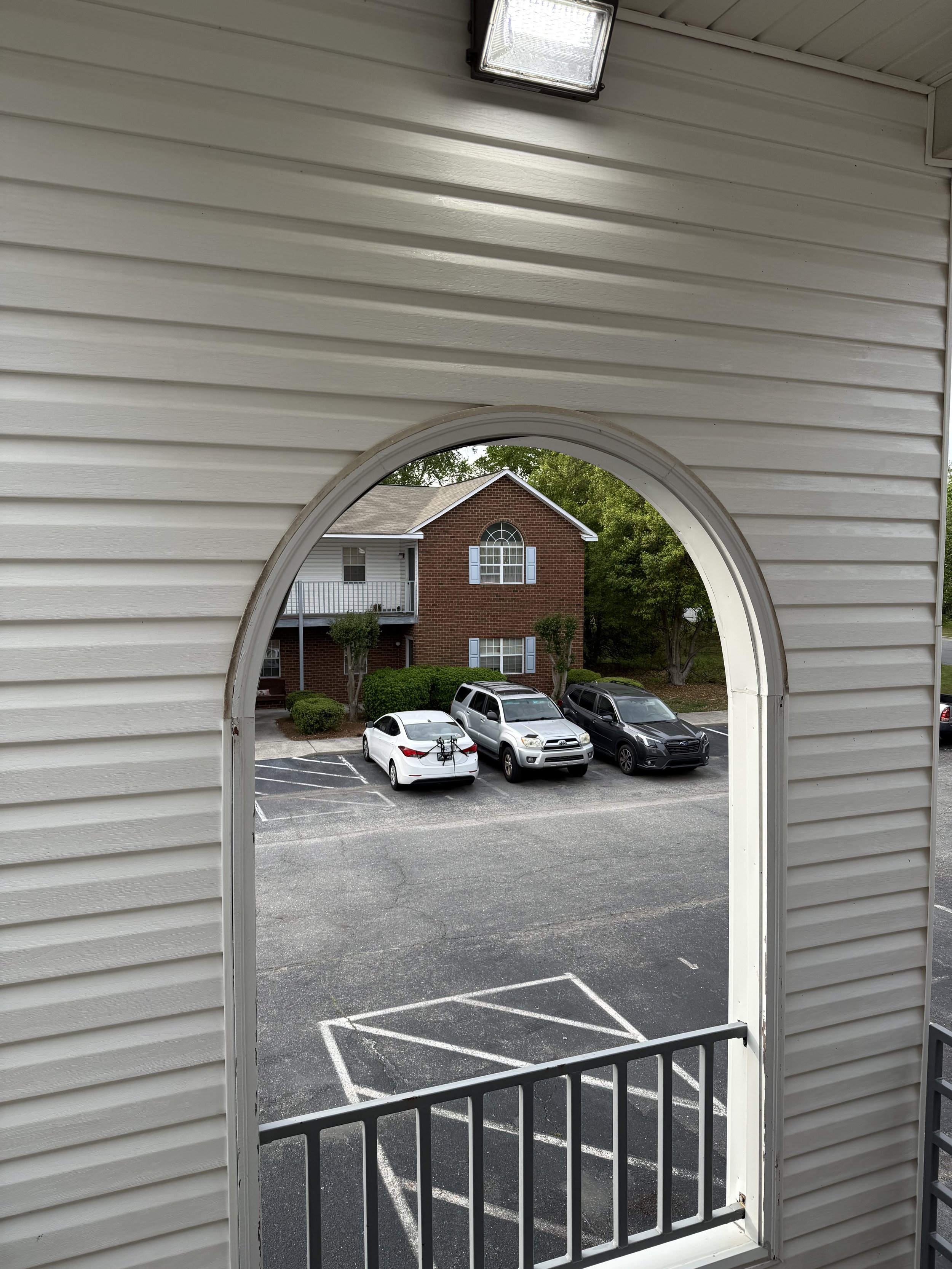 View through an arched window in a white house wall showing a parking lot with three cars, a grassy area with trees, and a red brick apartment building with blue shutters and a balcony.