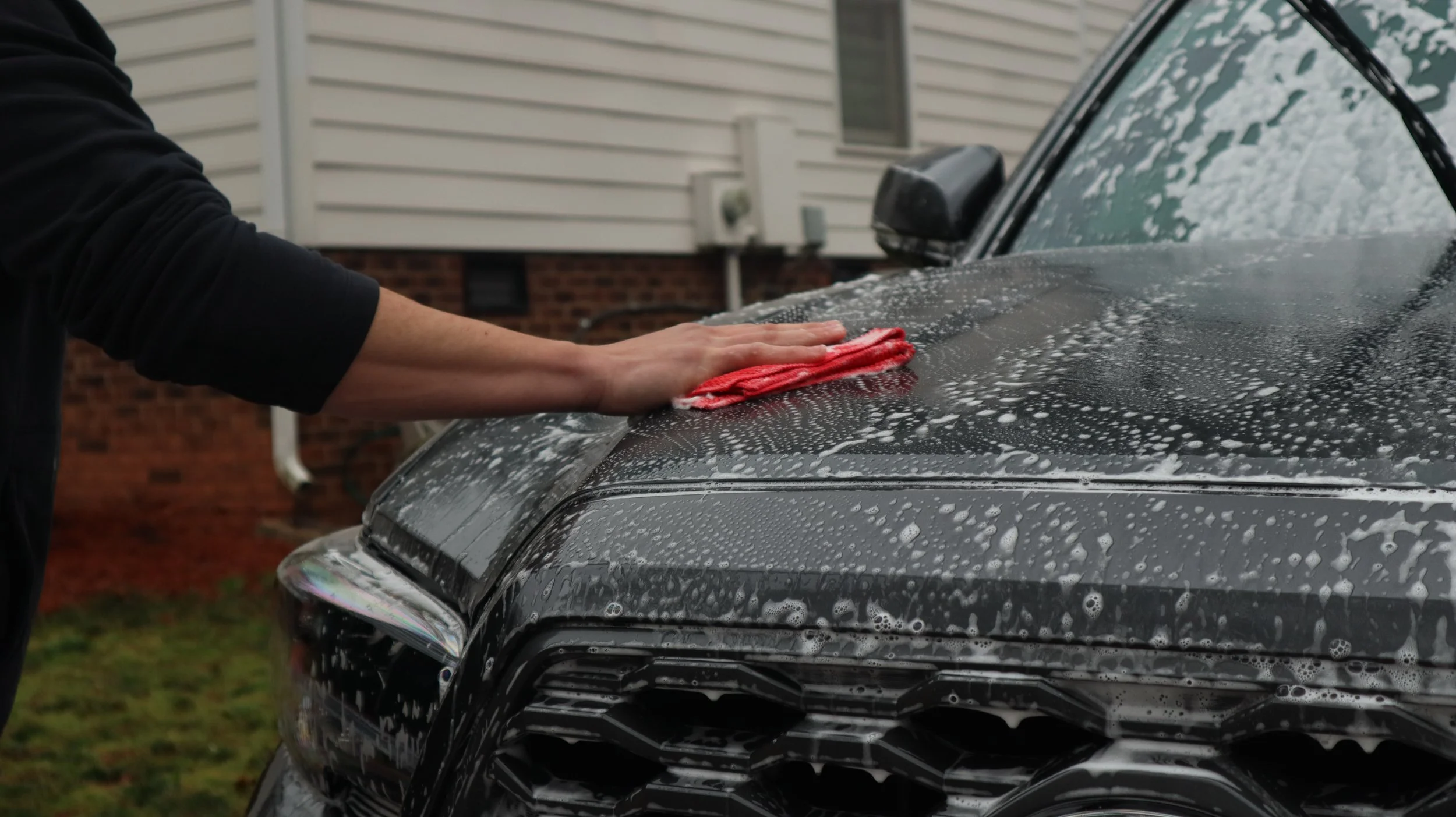 Person cleaning a black car with a red cloth, soap suds on the car, in a residential area with a house in the background.