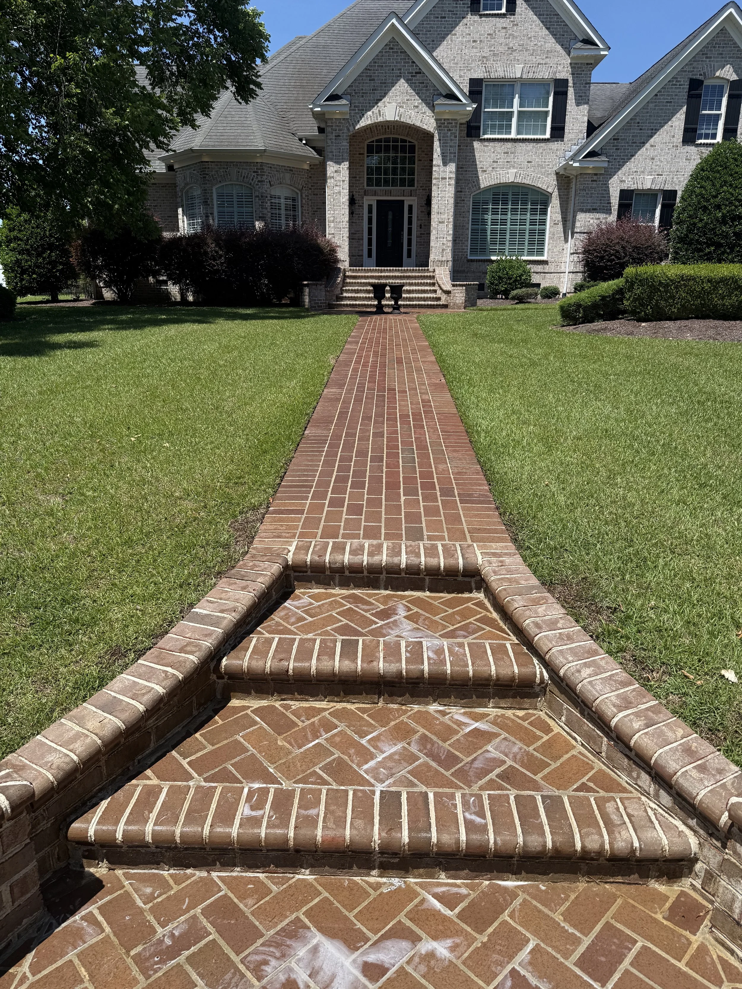 Brick pathway leading to the front steps of a large brick house, surrounded by green lawn and shrubbery.