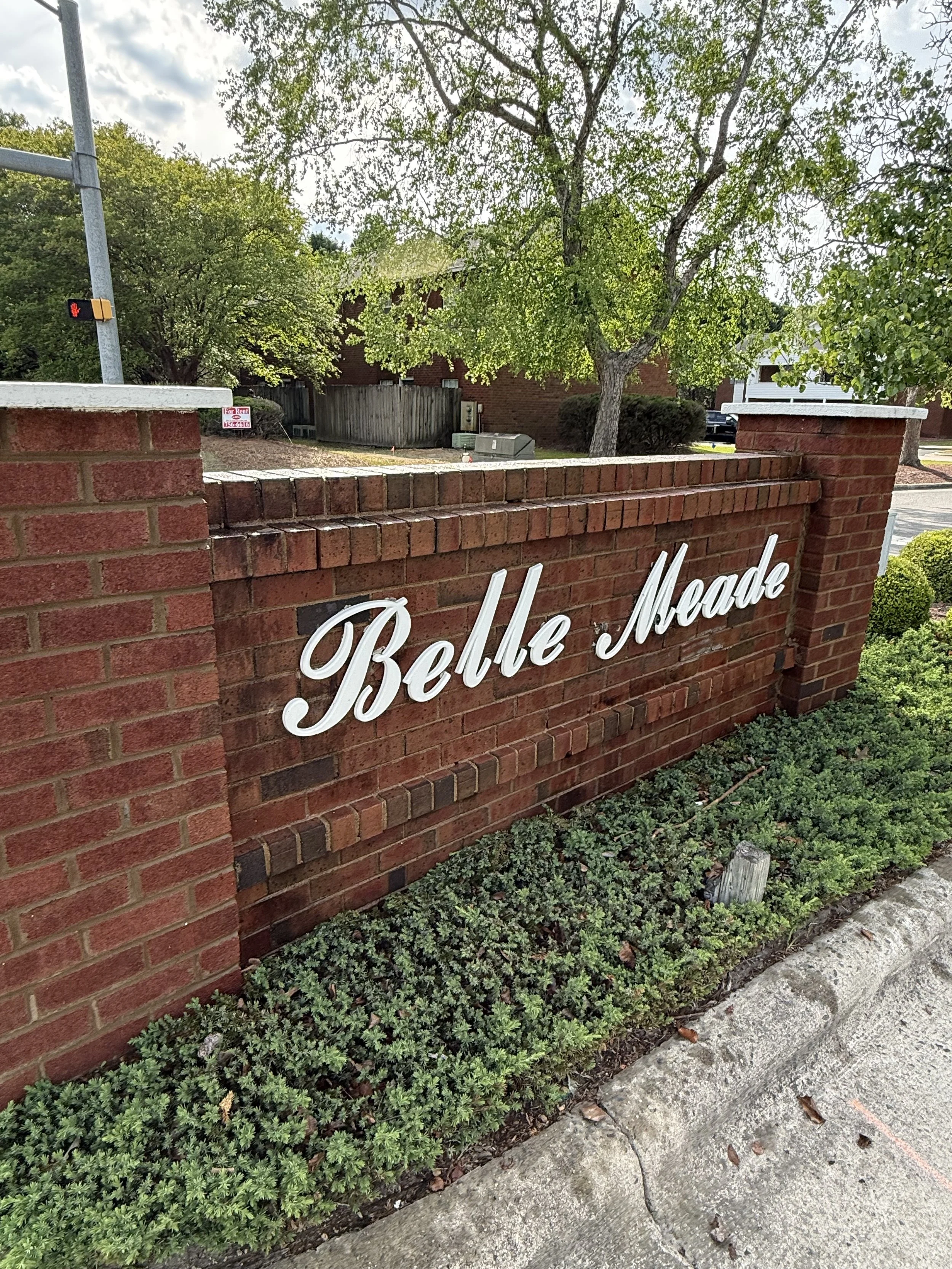 Brick wall with white cursive sign reading 'Piedmont' in front of trees and shrubs, with a sidewalk in the foreground.