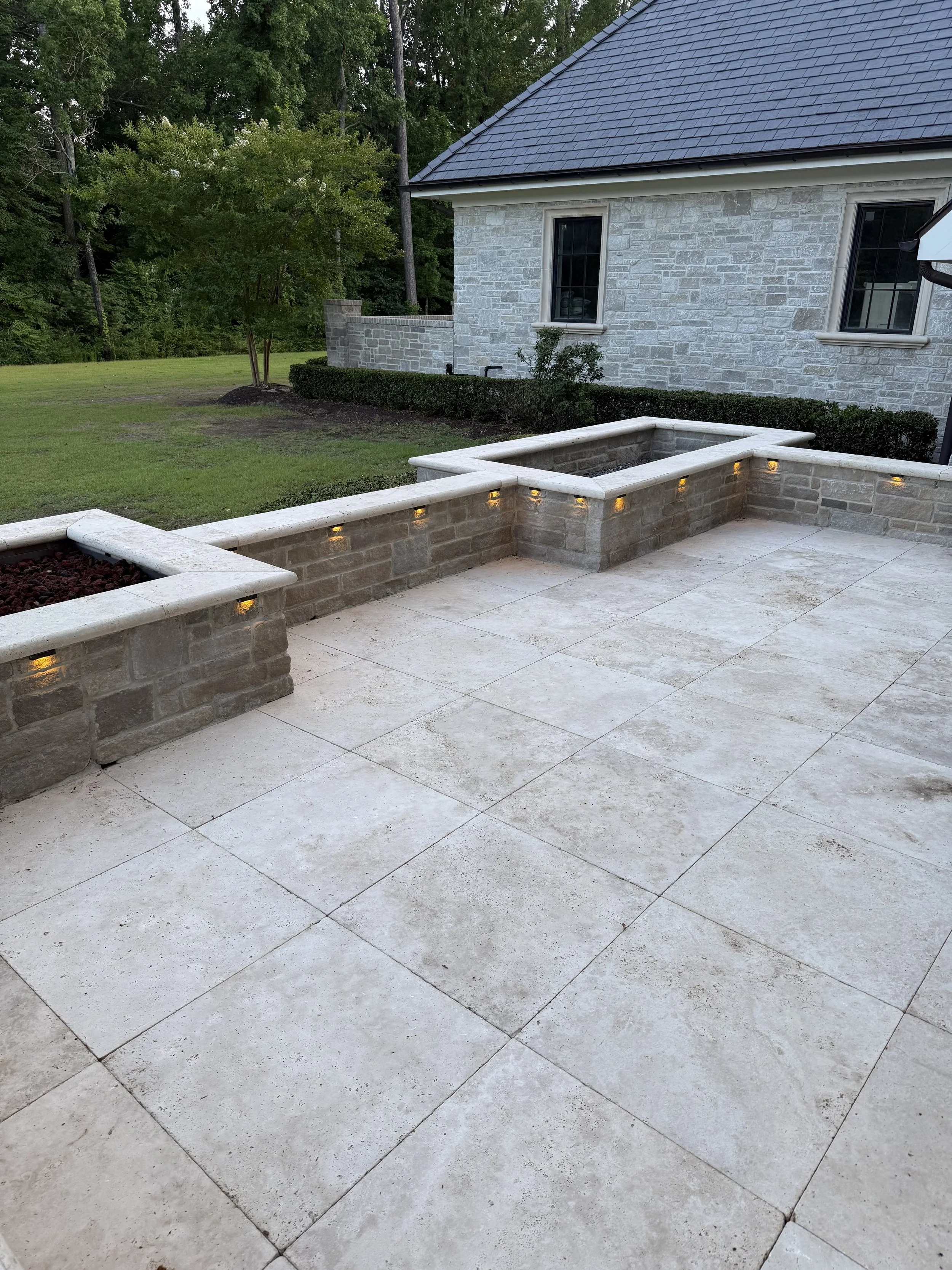 A backyard patio with large stone tiles, built-in stone planters with lighting, adjacent to a manicured lawn with a small tree, and a house with a stone exterior and a dark shingled roof.