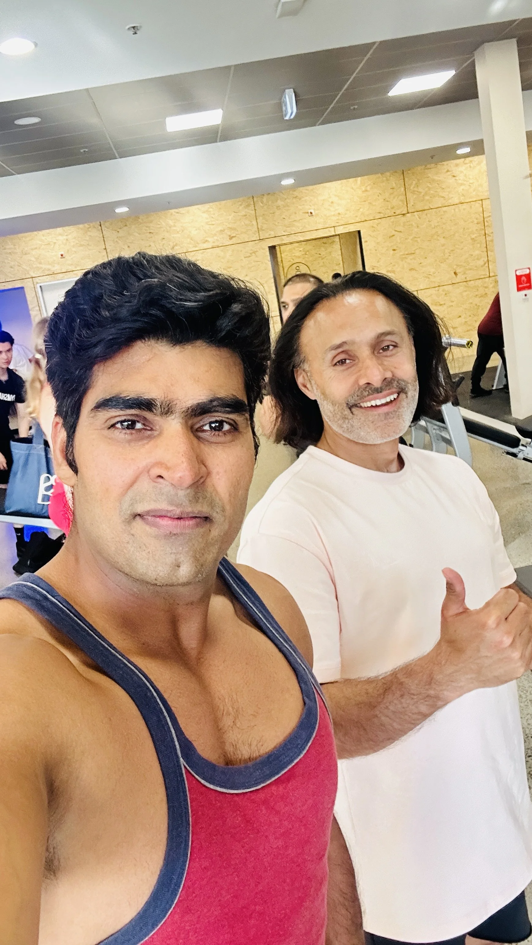 Two men taking a selfie at a gym. One is wearing a red tank top and the other a white t-shirt, giving a thumbs up. Several other people are visible in the background near gym equipment.