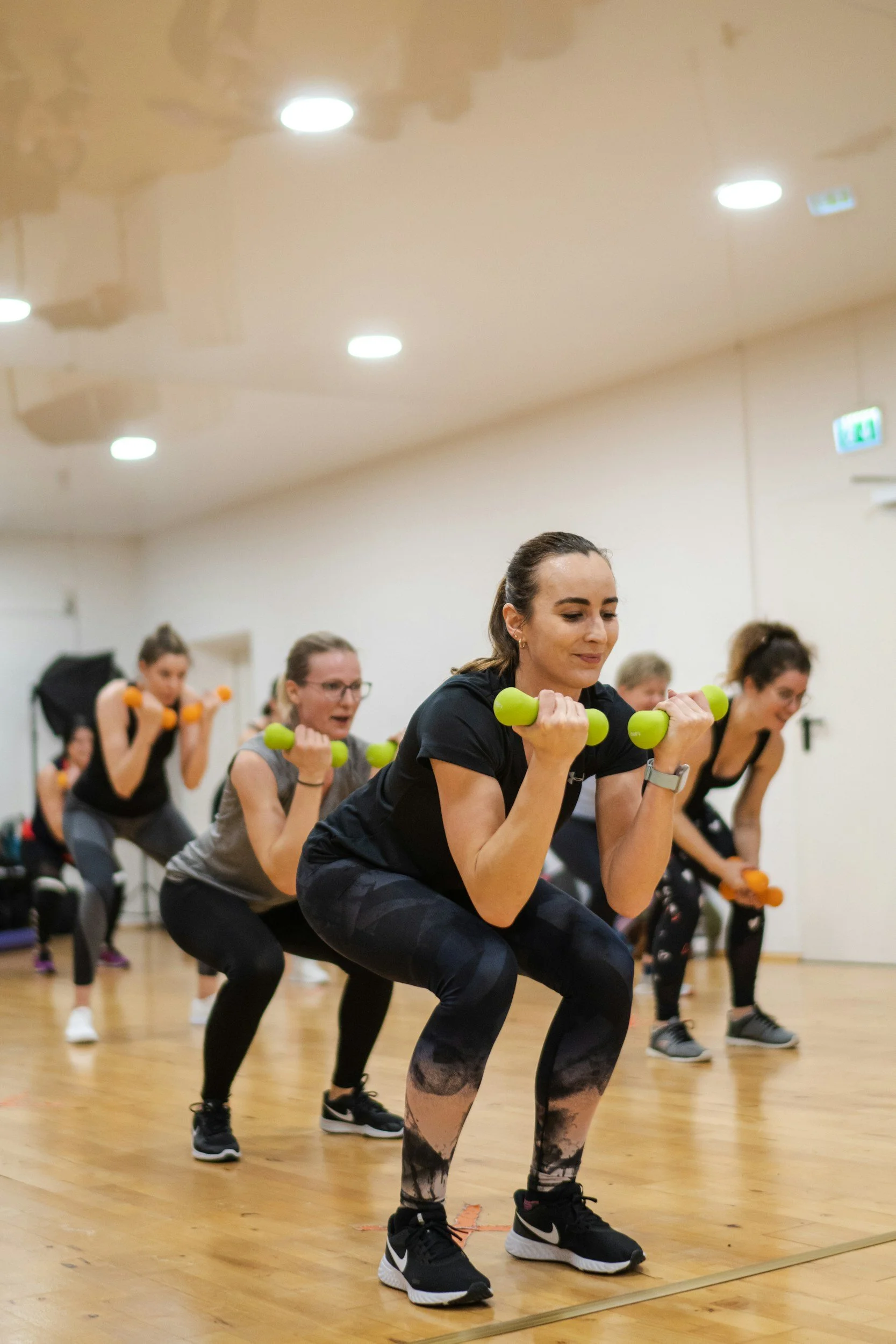 Women participating in a fitness class, performing squats with small green dumbbells in a spacious room with wooden floor and ceiling lights.