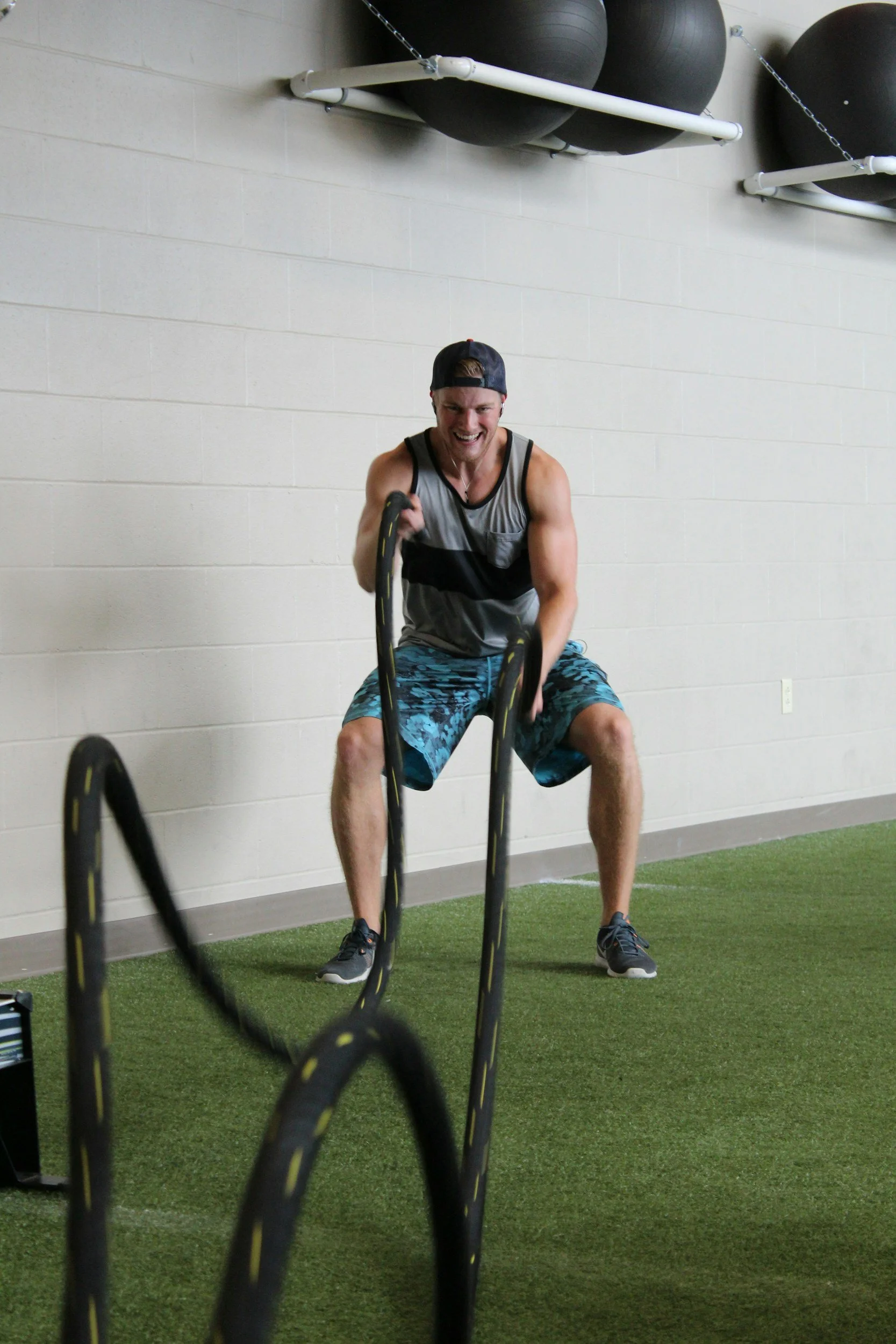 A young man exercising with battle ropes in a gym, smiling and wearing a gray tank top, blue shorts, gray sneakers, and a backward cap. The gym has a white brick wall, black exercise balls on racks, and green turf flooring.