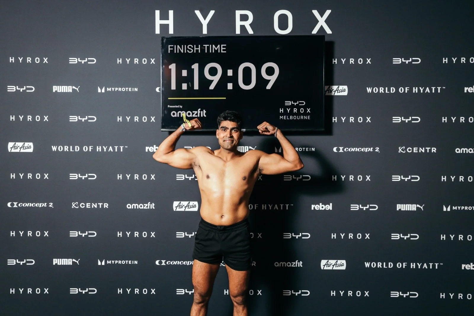 A shirtless man with black shorts standing in front of a black backdrop, flexing his biceps, with a large screen behind him showing a finish time of 1:19:09 and various sponsor logos.