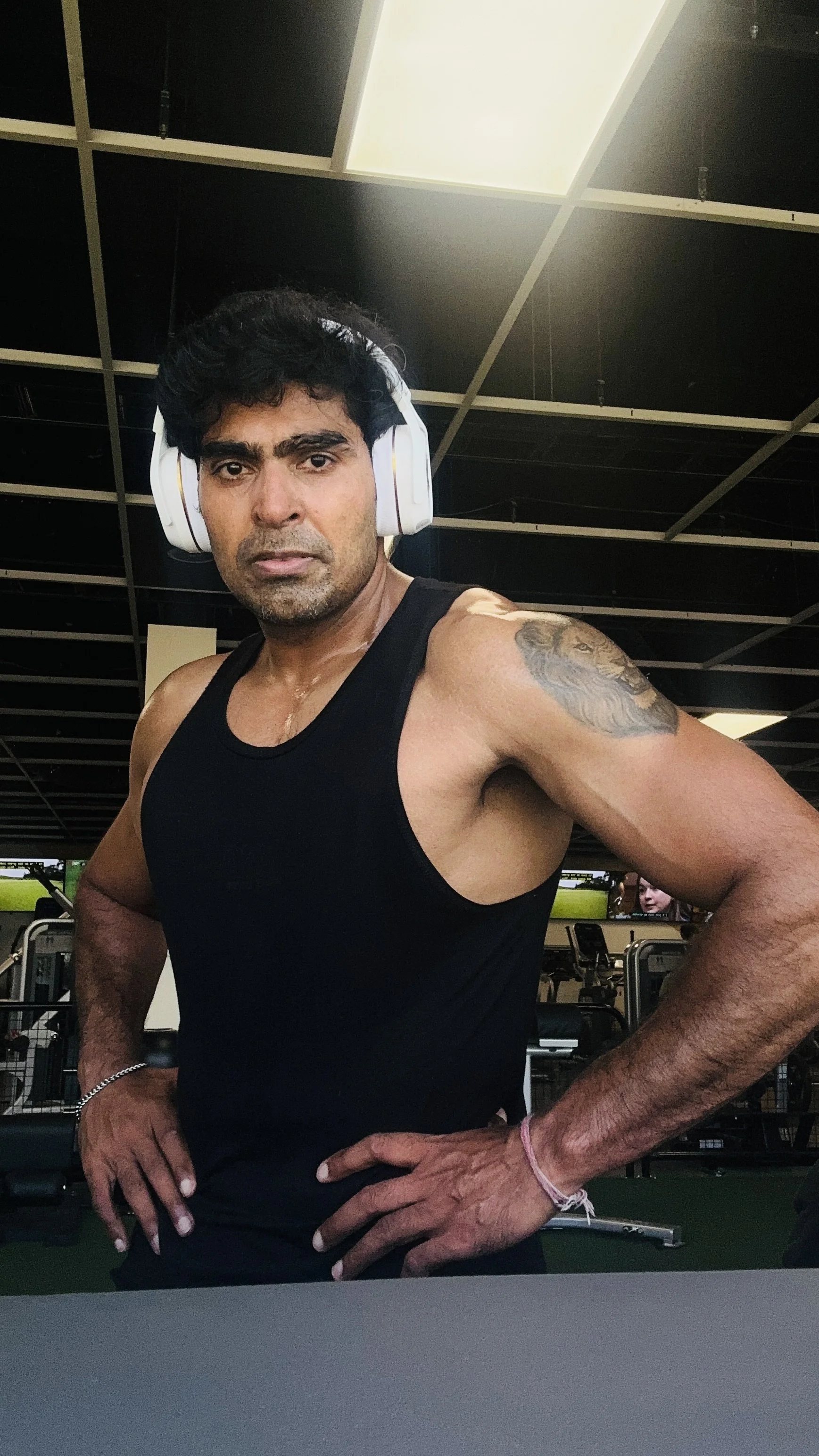 A man wearing a black tank top and large white headphones standing in a gym with his hands on his hips, with a black and white ceiling and fitness equipment in the background.