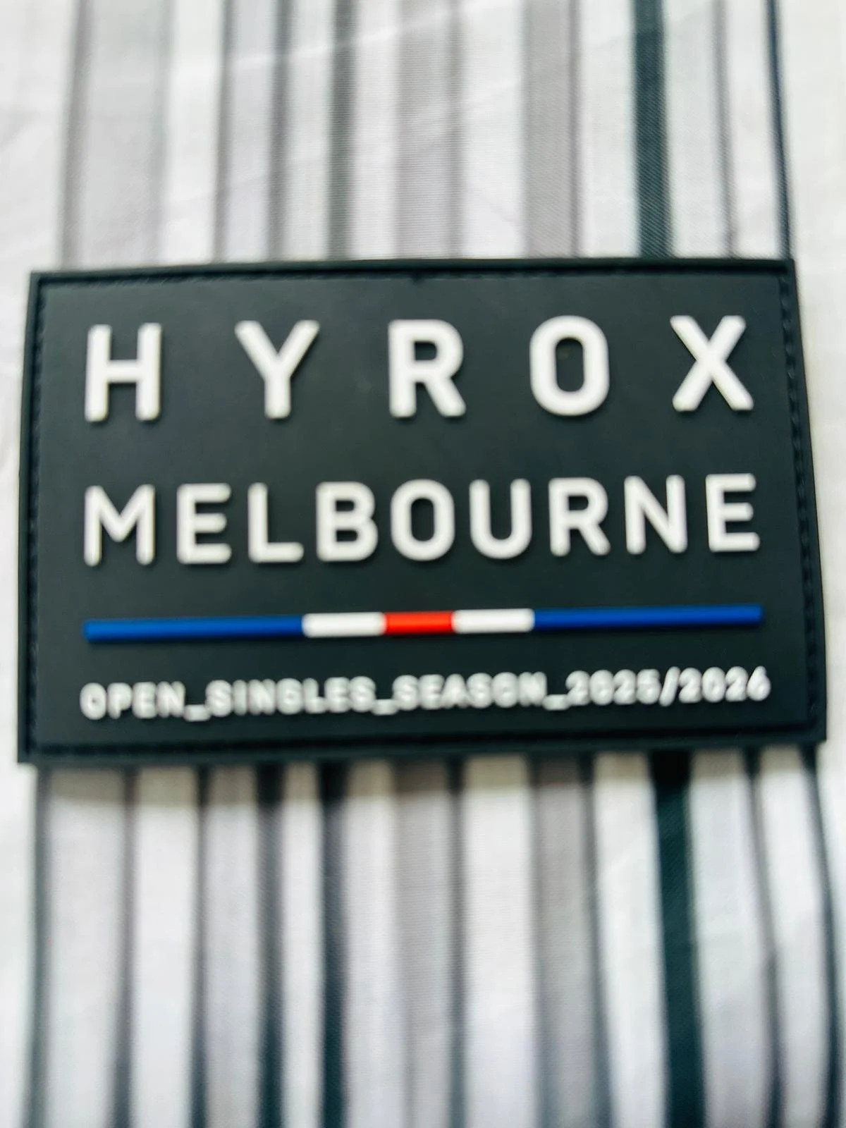 Black sign with white letters displaying "HYROX MELBOURNE," flanked by three horizontal stripes in blue, white, red, white, and blue. Smaller white text below reads "OPEN_SINGLES_SEASON_2023/2026." The sign is attached to a metal fence, with a semi-t