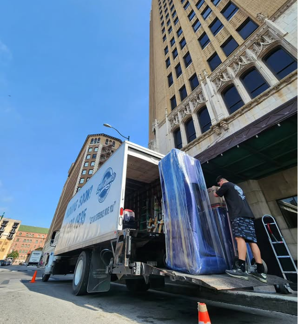 A person loading or unloading large glass panels from a moving truck on a city street, with tall buildings and blue sky in the background.