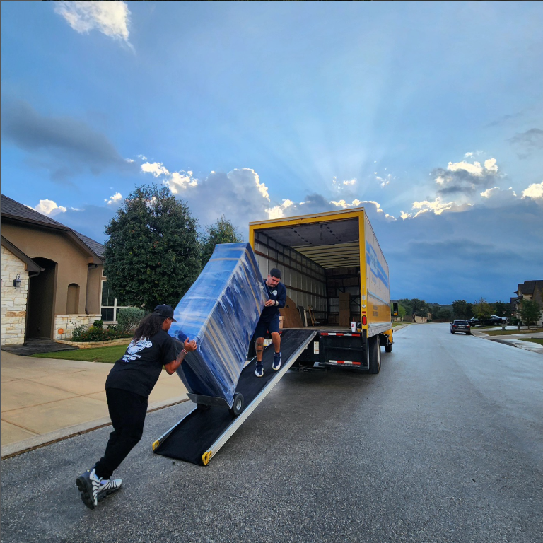 Two people loading a large blue and white object into a moving truck on a suburban street at sunset.