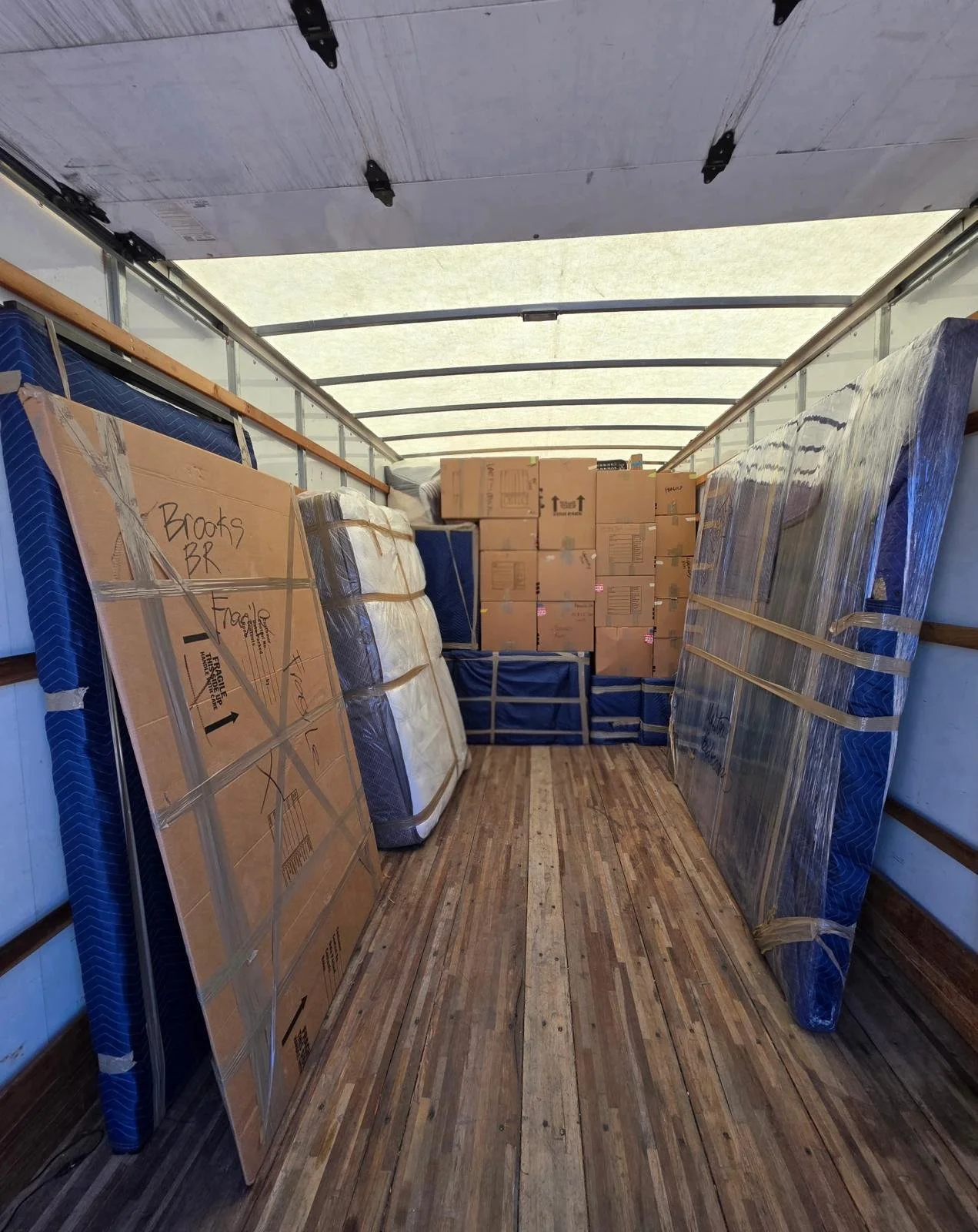 The image shows the inside of a moving truck filled with packed boxes and furniture, including mattresses wrapped in plastic, and labeled cardboard boxes stacked at the back. The wooden floor of the truck is visible, with a partly open rear door lett