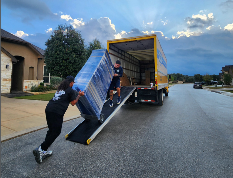 Two people unloading a large mattress from a moving truck on a residential street during the day.