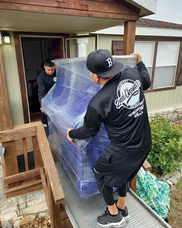 Two young men are moving a large, wrapped, blue glass or acrylic piece into a house through the front door. One man is wearing a black jacket with white graphics and a black baseball cap, while the other is in a dark shirt.