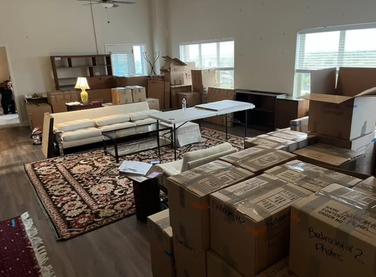Living room filled with numerous packed and unpacked moving boxes, some stacked on each other, with a white sofa, a coffee table, a small side table, and a patterned area rug. The room has large windows with blinds and minimal furniture.