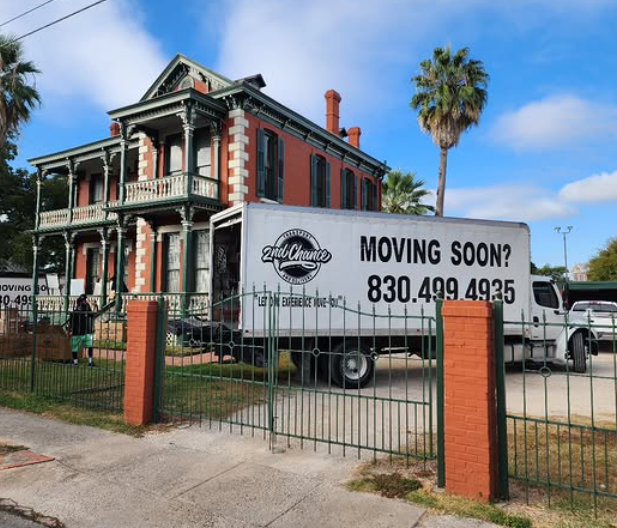 A large moving truck parked in front of a historic, two-story, red brick house with a porch and black shutters. The truck has a sign that reads 'MOVING SOON?' and a phone number. The house is surrounded by a black wrought iron fence, with palm trees 