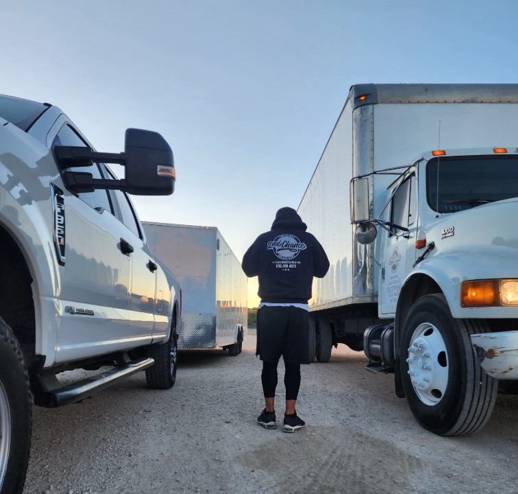 A person standing between a white pickup truck on the left and a white semi-truck on the right, outdoors during sunset or sunrise.