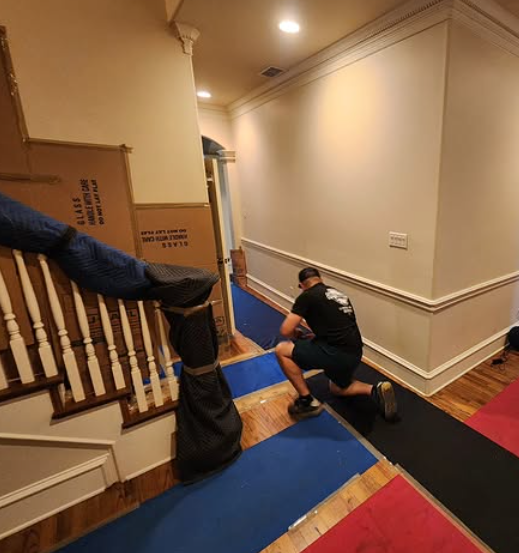 A person is kneeling on the floor, installing or fixing a blue carpet runner at the bottom of a staircase in a hallway, with a large cardboard box leaning against the staircase railing nearby.