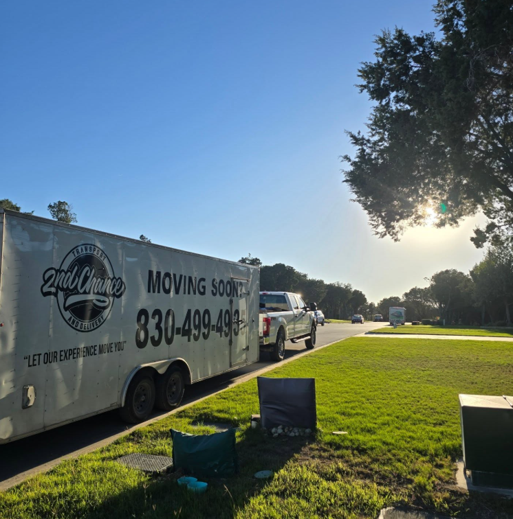 A moving truck and several cars on a suburban street during sunset. The truck has the text '2nd Chance Transport' and 'MOVING SOON' along with a phone number. There is a grassy area with some bags and small objects near the sidewalk.