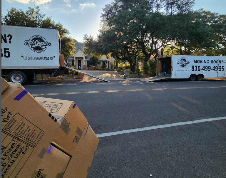 Moving trucks and cardboard boxes on a residential street with a house and large trees in the background.