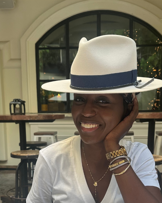 A woman smiling while wearing a large fedora hat, sitting in a cozy restaurant or cafe with a large arched window in the background.