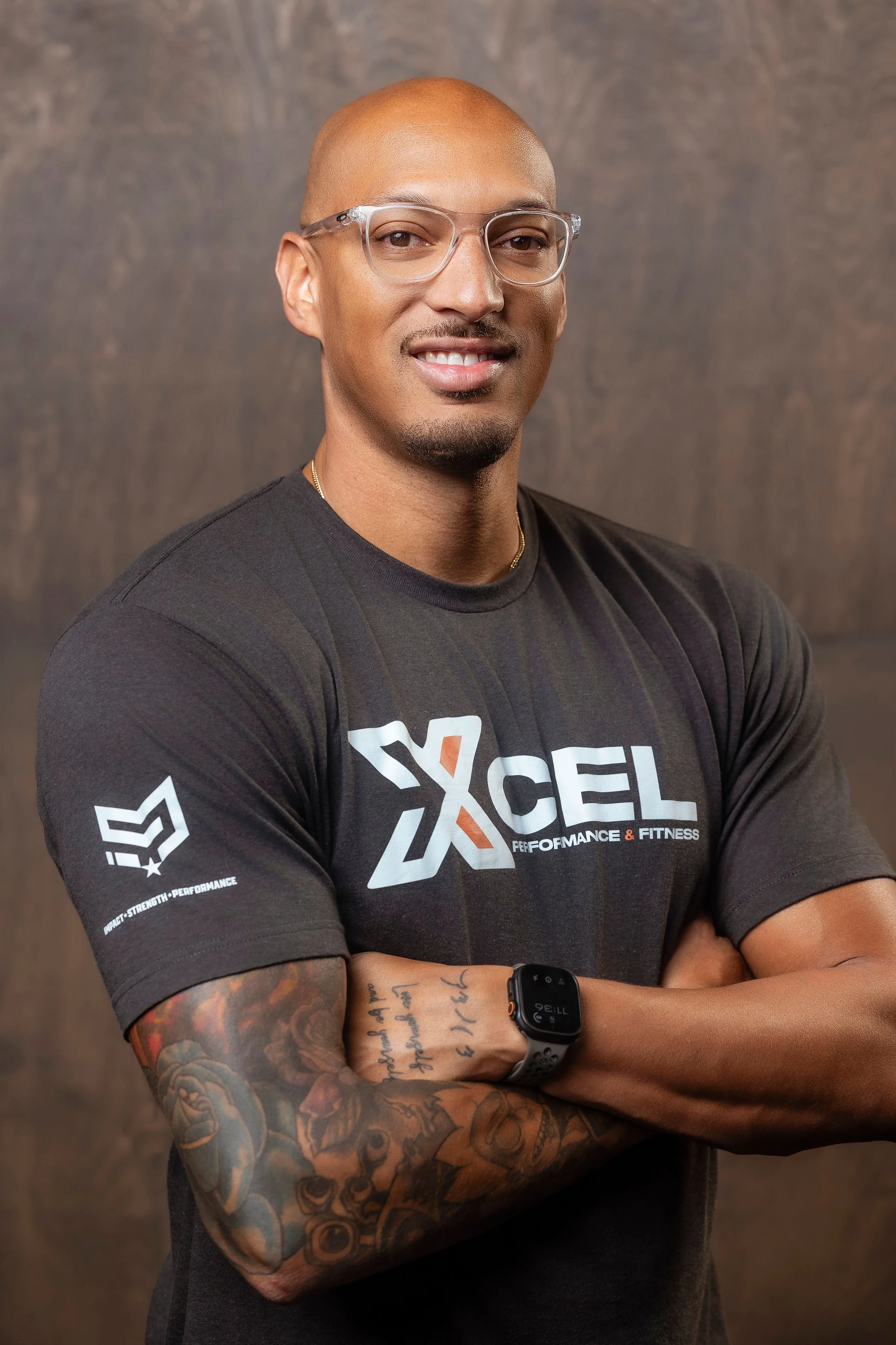 A man with tattoos on his arm, wearing glasses, a black fitness T-shirt, and a black fitness watch, standing with his arms crossed and smiling in front of a brown textured background.