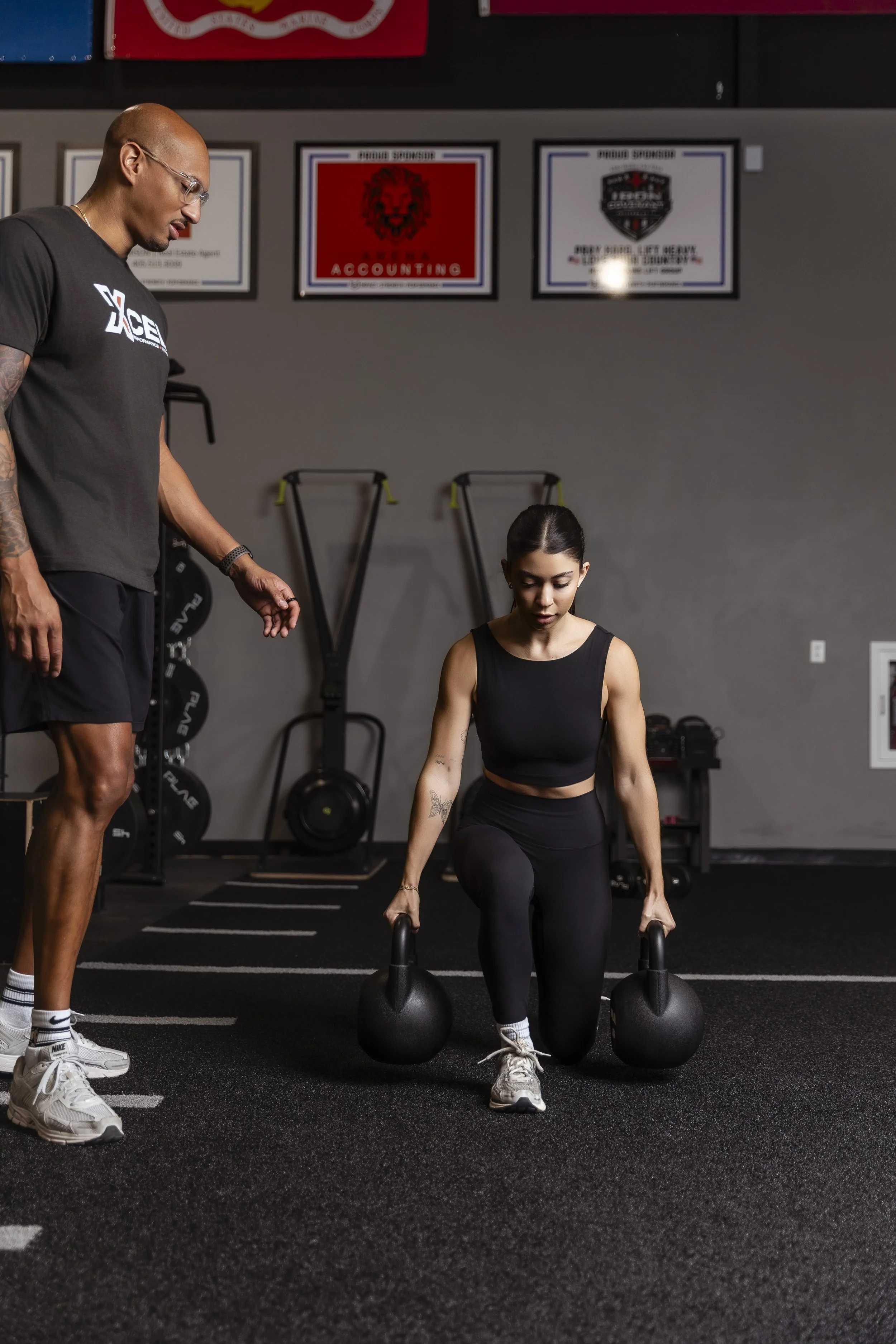 A woman in black athletic clothing lifting two large kettlebells in a gym, with a trainer standing nearby observing, against a background of gym equipment and framed posters on the wall.
