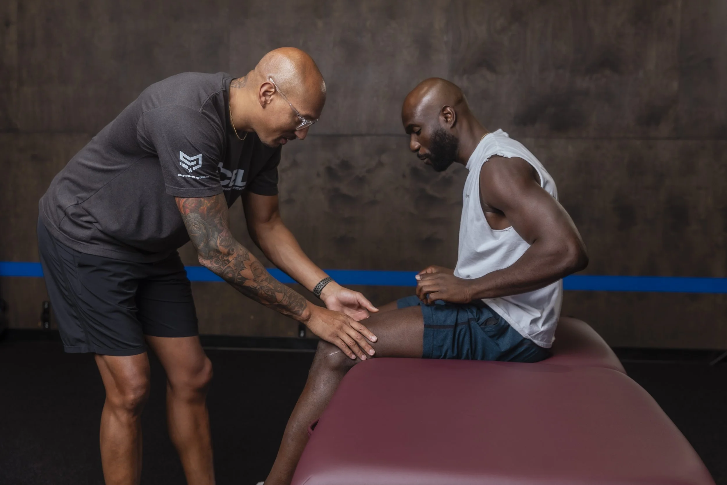 A person is sitting on a treatment table with their leg extended, while another person appears to be a trainer or therapist assisting with leg exercises or rehabilitation in a gym or physical therapy setting.