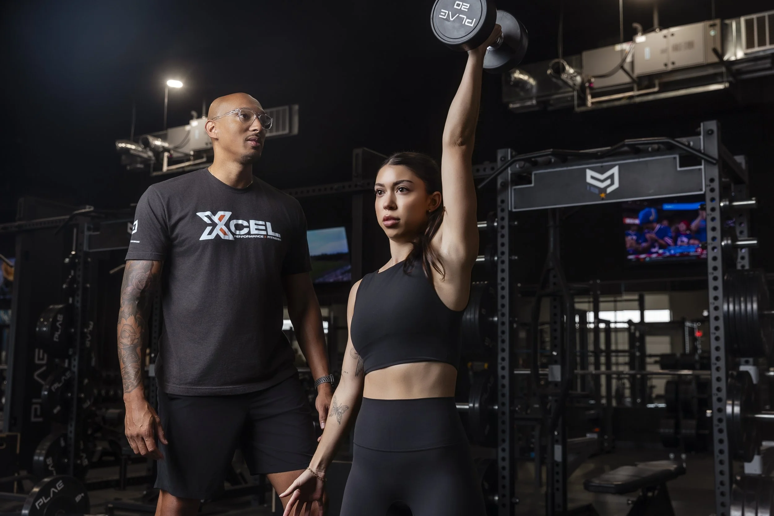 A young woman in workout clothes lifting a dumbbell while a strength coach observes in a gym.