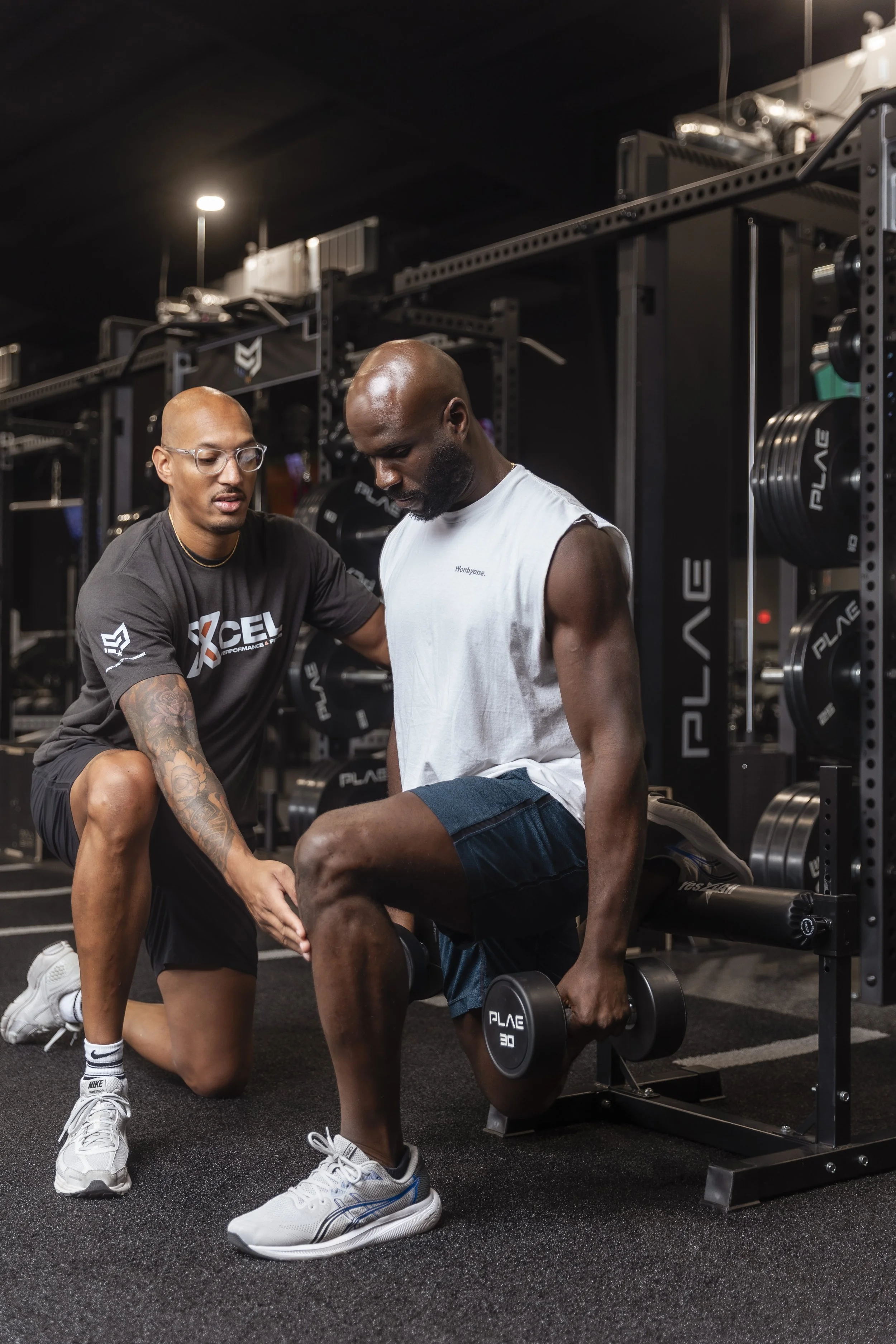 A physical therapist assisting a man with a dumbbell exercise in a gym. The physical therapist is kneeling and guiding the man, who is holding a dumbbell, on a workout bench. The gym has weight racks with plates and other equipment in the background.