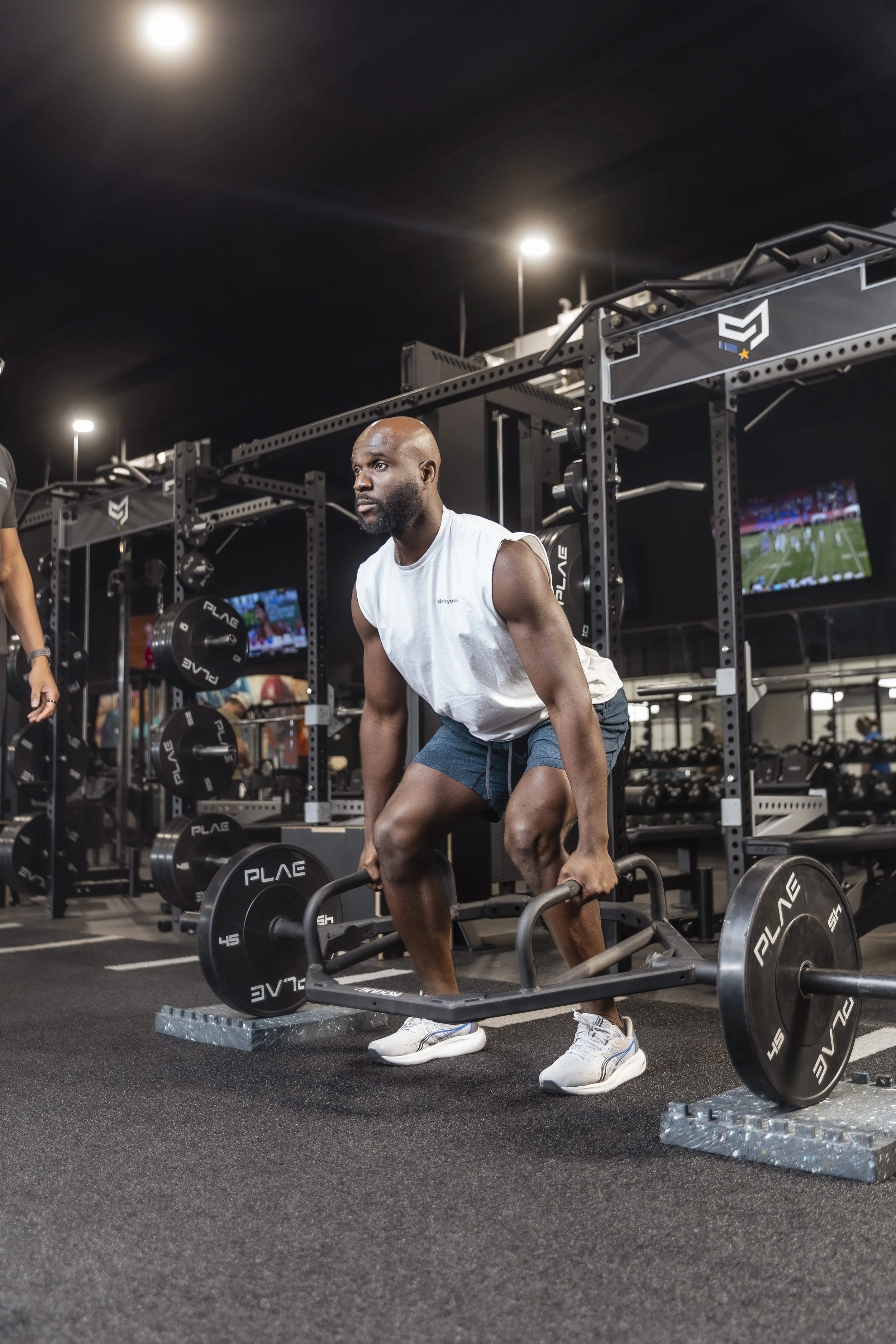 Man lifting weights in a gym, using a trap bar with weights, with gym equipment and TV screens in the background.