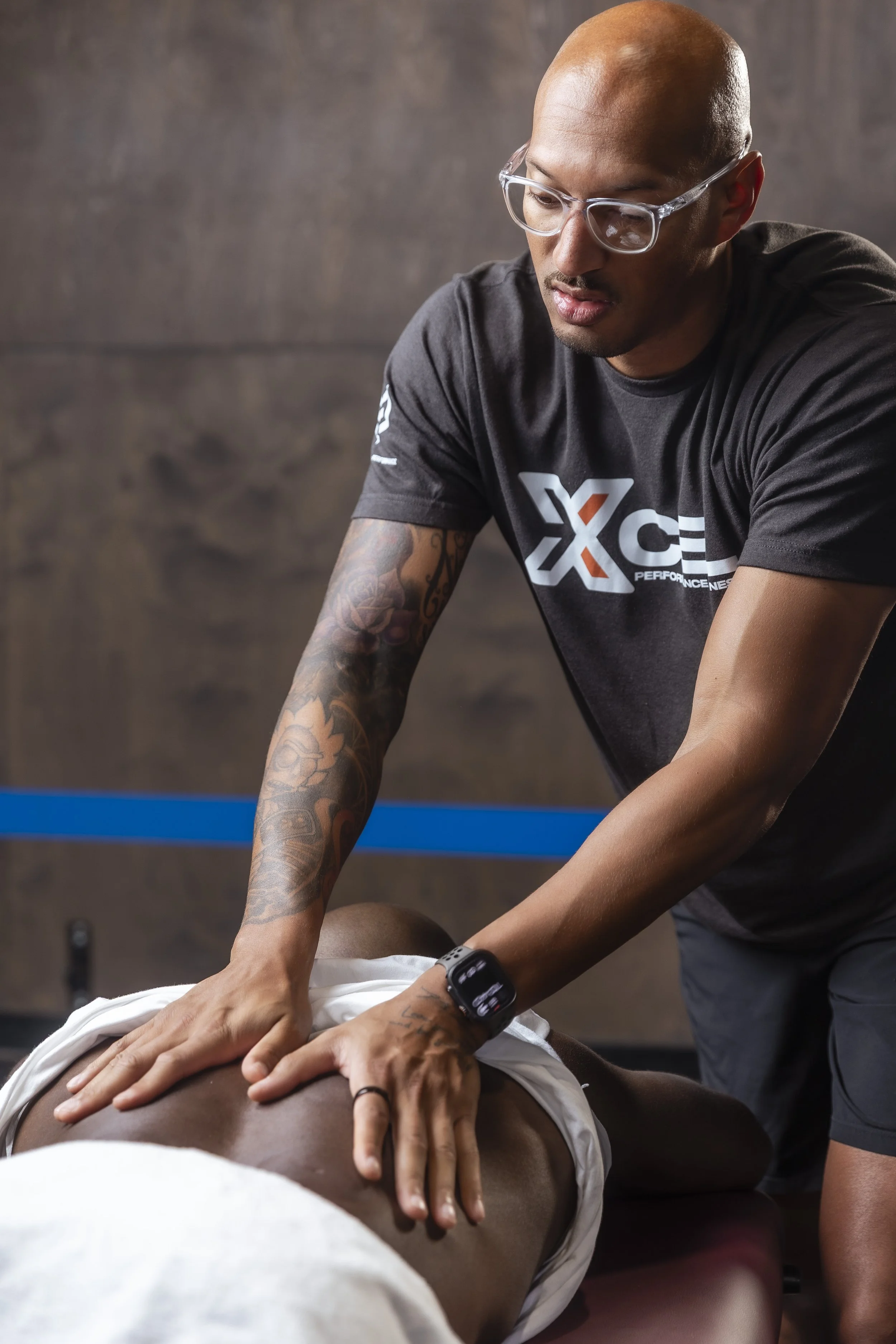 A person receiving a massage from a physical therapist in a gym or wellness center. The therapist is wearing a black t-shirt with a logo and glasses, while the client is lying face down on a massage table with hands resting on their back.