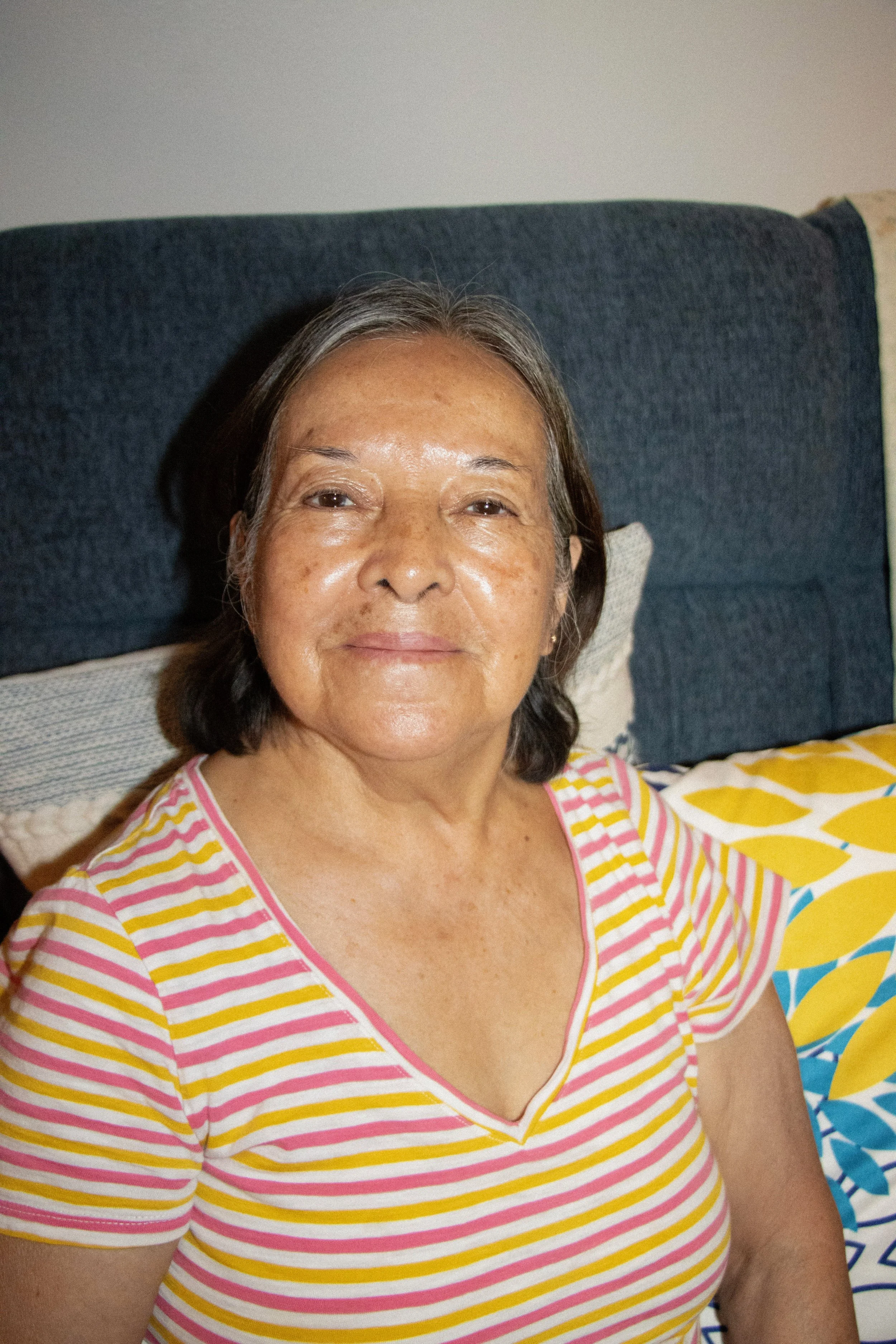 A woman with gray hair and a warm smile, wearing a short-sleeved, yellow and pink striped shirt, seated on a bed with a dark headboard and colorful bedding.
