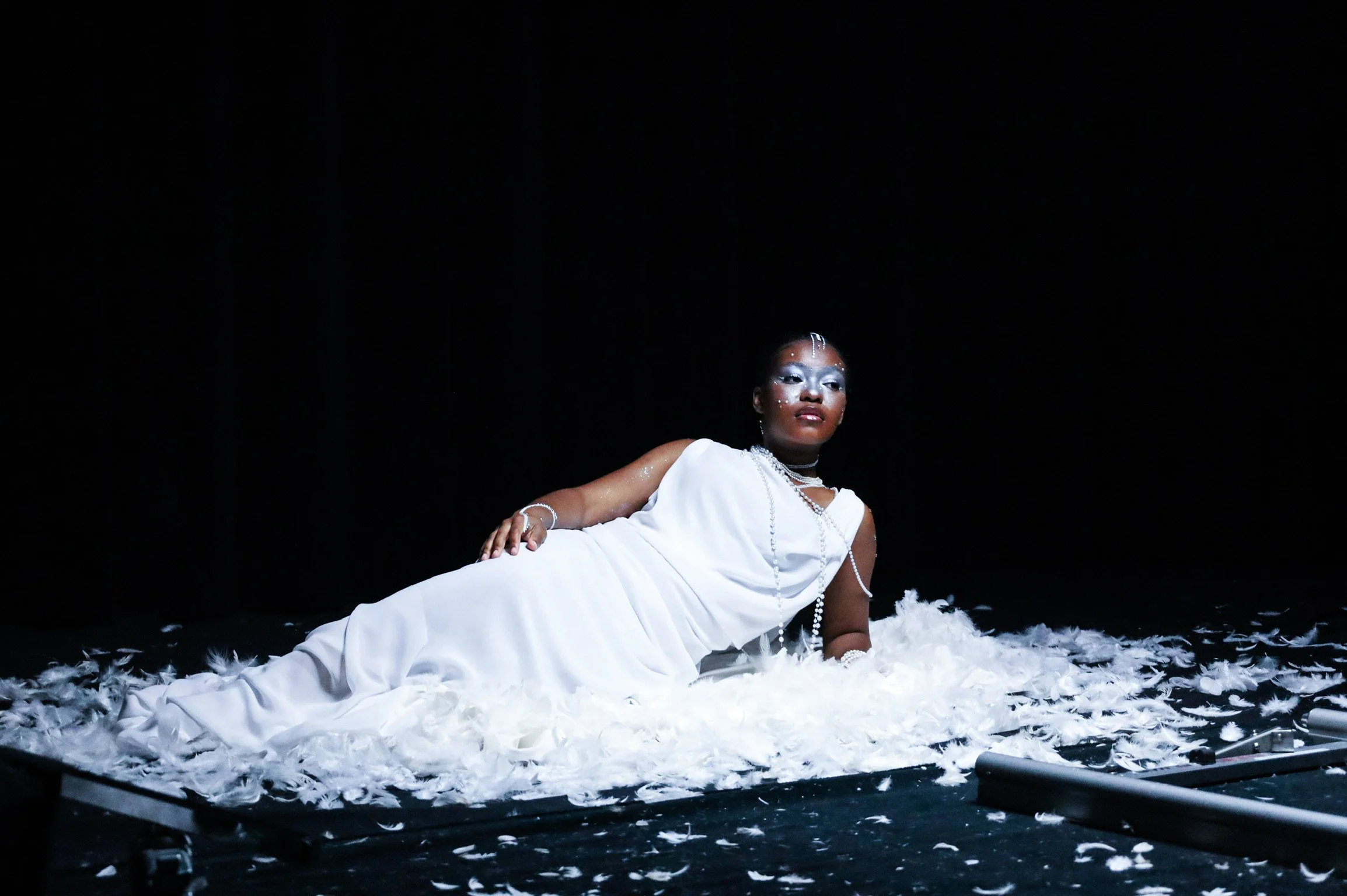 A performer dressed in white lying on a stage covered with white feathers, with a dark background.