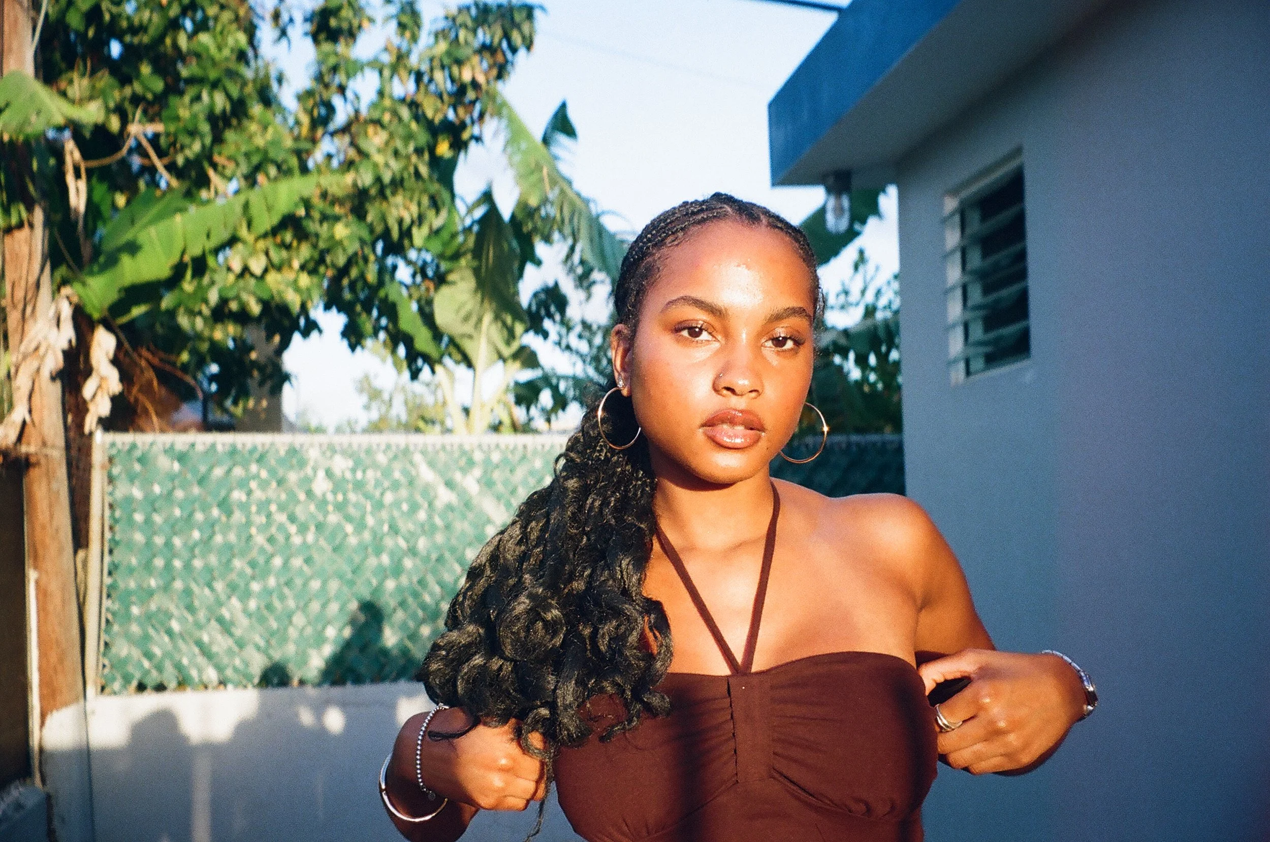 A young woman with braided hair and hoop earrings standing outdoors on a sunny day. She is wearing a strapless brown top and jewelry, with a background of green trees, a blue sky, and part of a house.