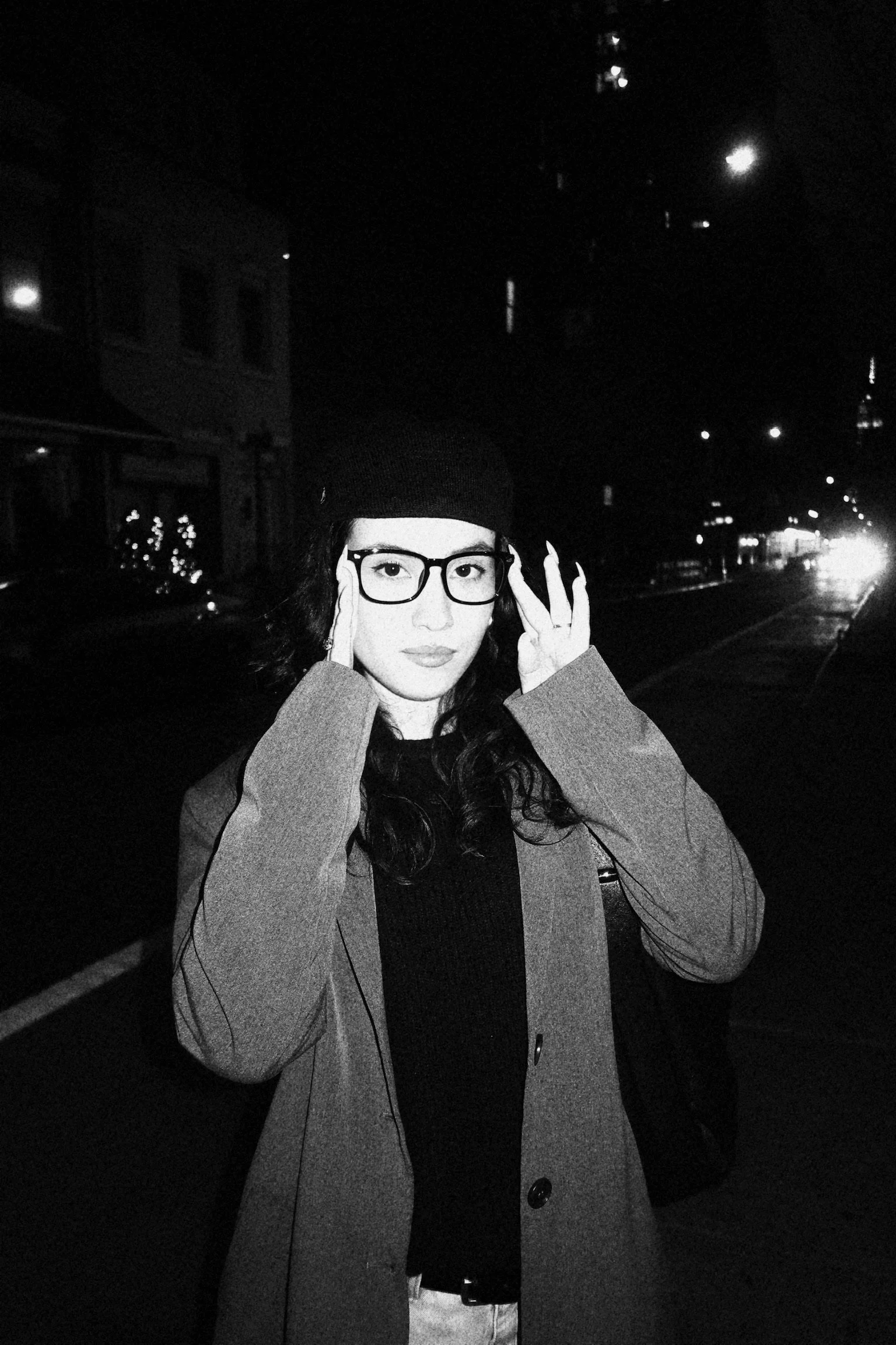 A woman wearing glasses and a black beanie stands outdoors at night in a city street, holding her glasses with both hands.