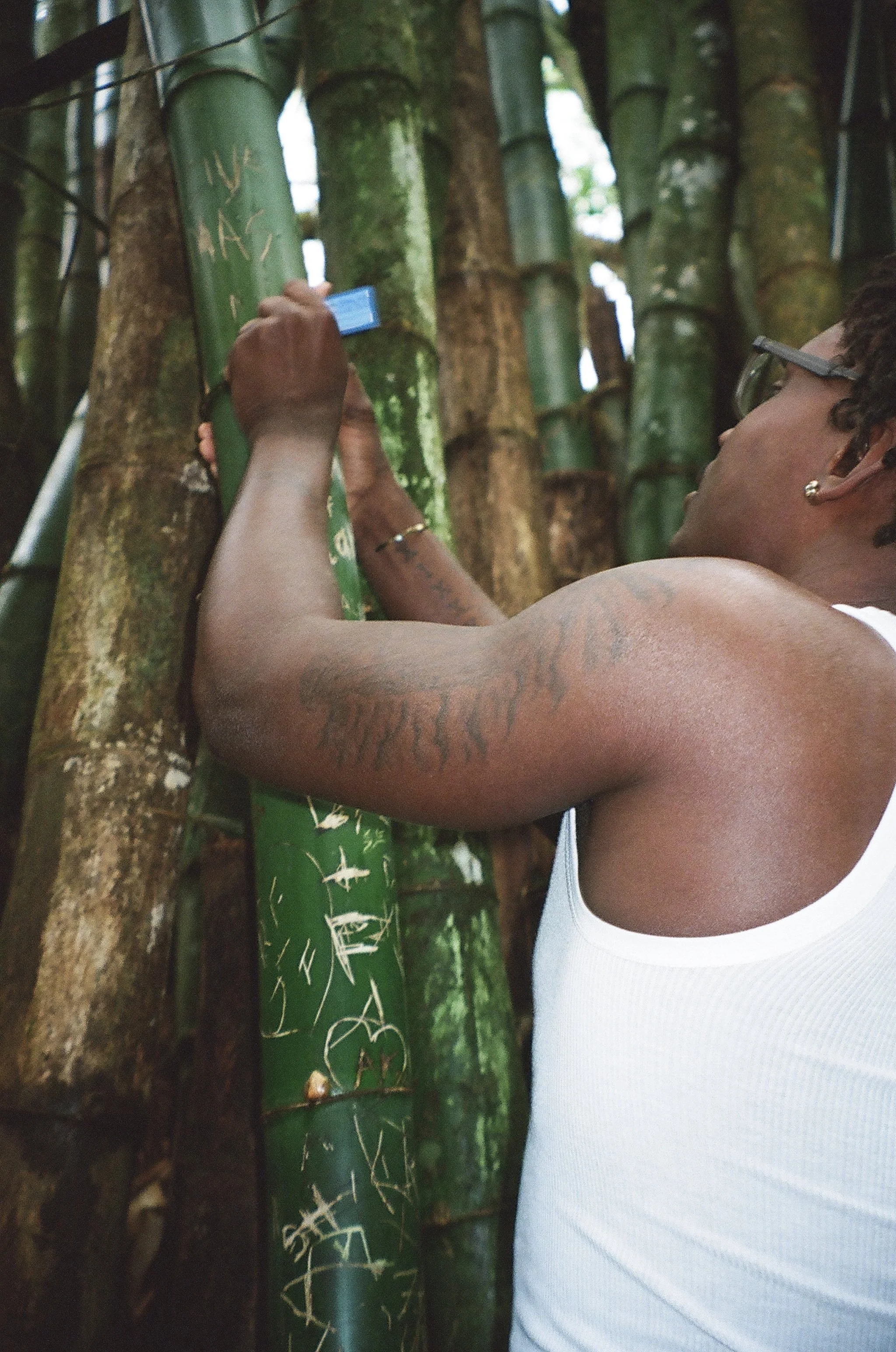 A woman with curly hair and glasses wearing a white tank top is writing on a green bamboo stalk with white chalk in a forest.