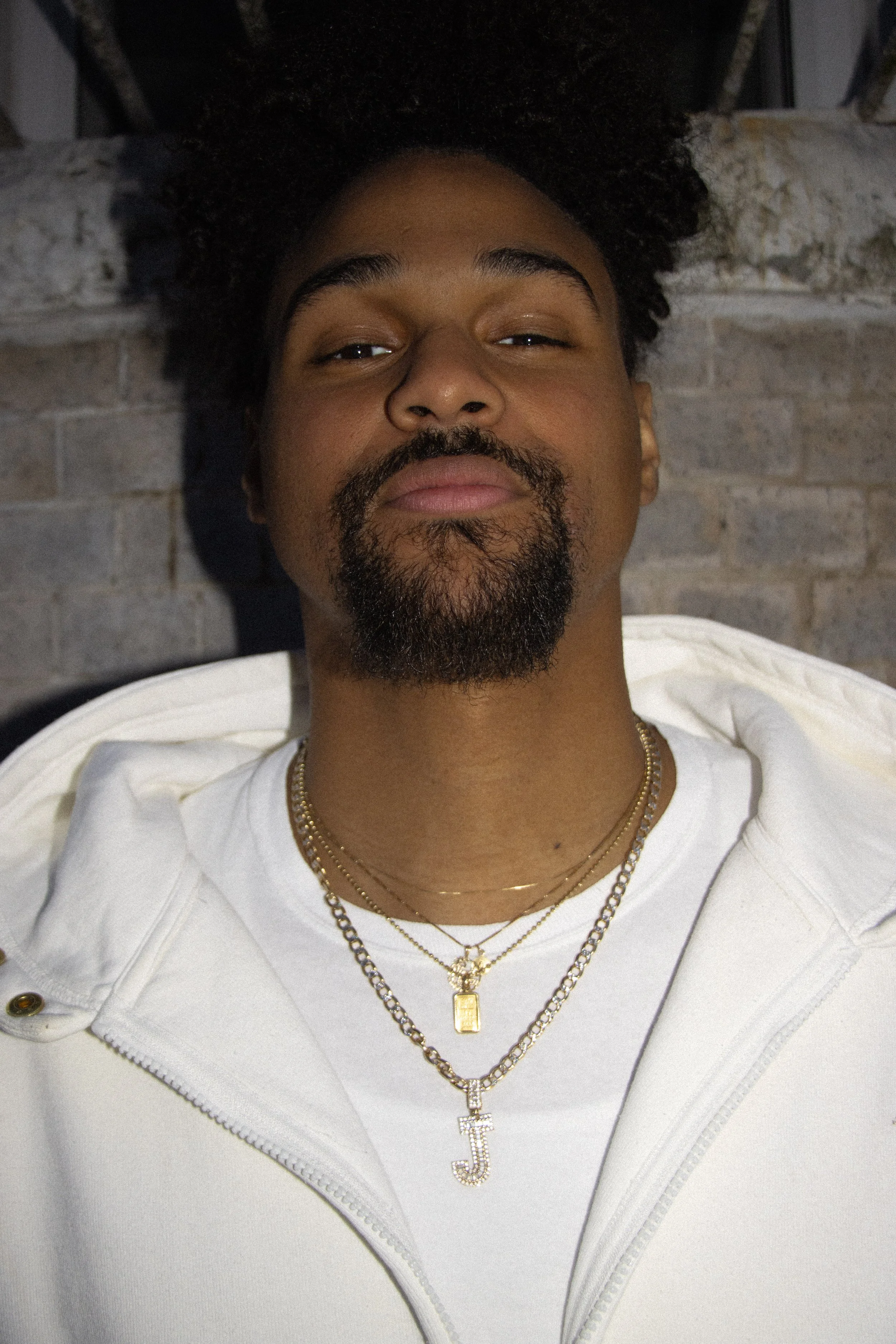 Close-up of a man with curly hair and a beard, wearing layered gold necklaces with letter charms, a white zip-up hoodie, and standing in front of a brick wall.