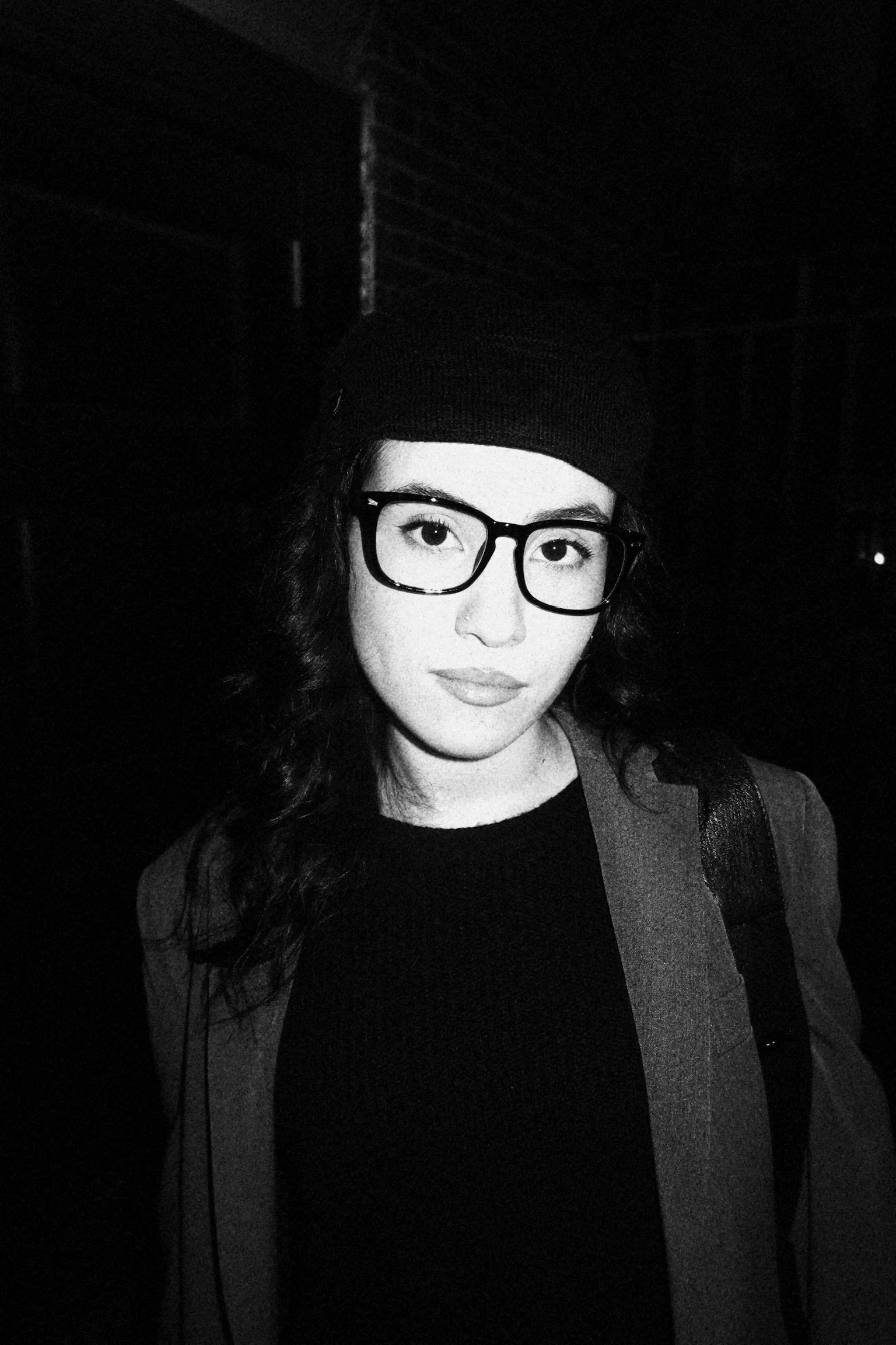 A black-and-white photo of a young woman with dark hair, wearing glasses, a beanie, and a jacket, standing in a dark environment.
