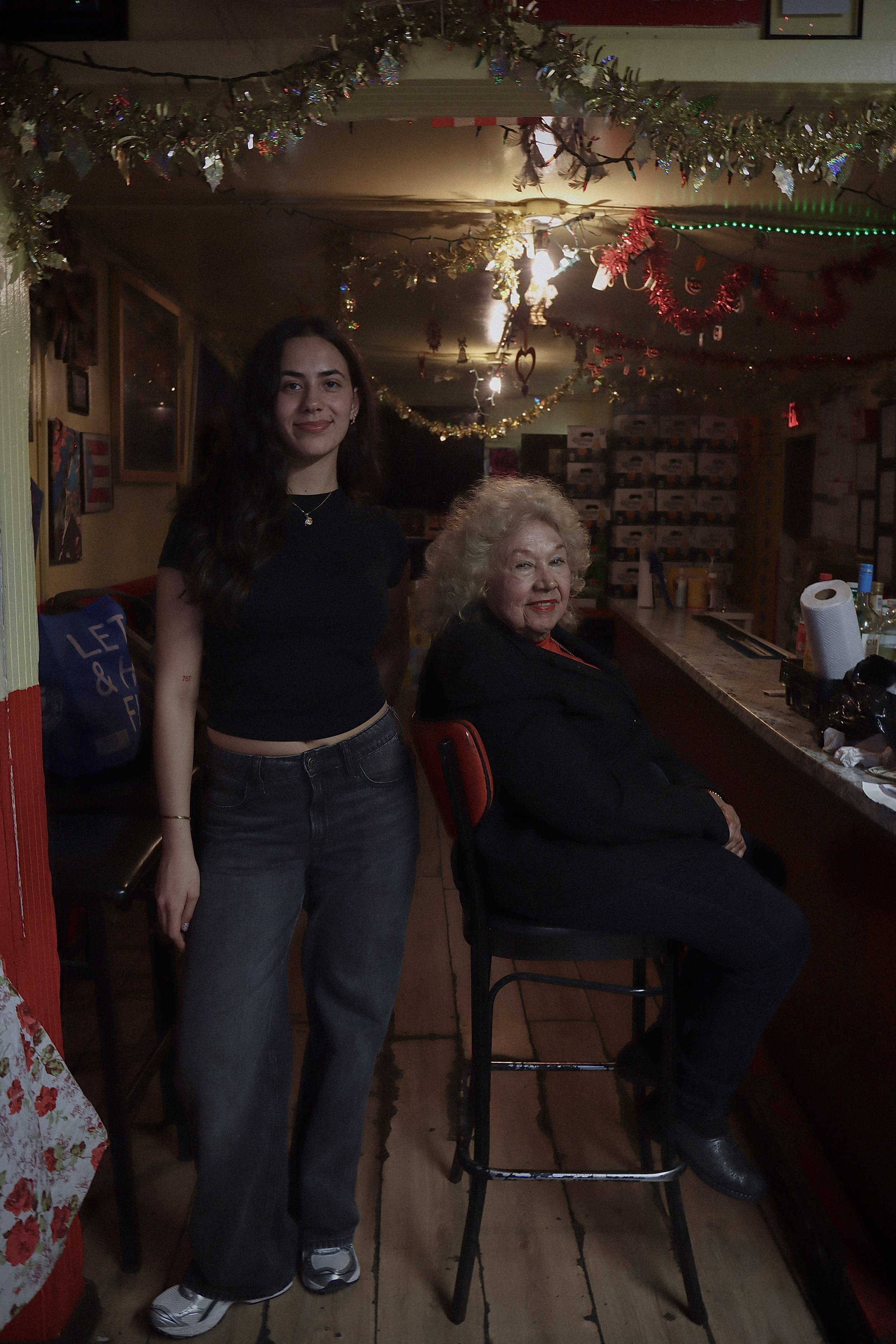 A young woman with long dark hair, wearing a black t-shirt and jeans, standing next to an older woman with curly gray hair, wearing a black top, sitting on a barstool. The scene is decorated with Christmas tinsel and ornaments hanging from the ceiling, and there are various items on the bar counter behind them.