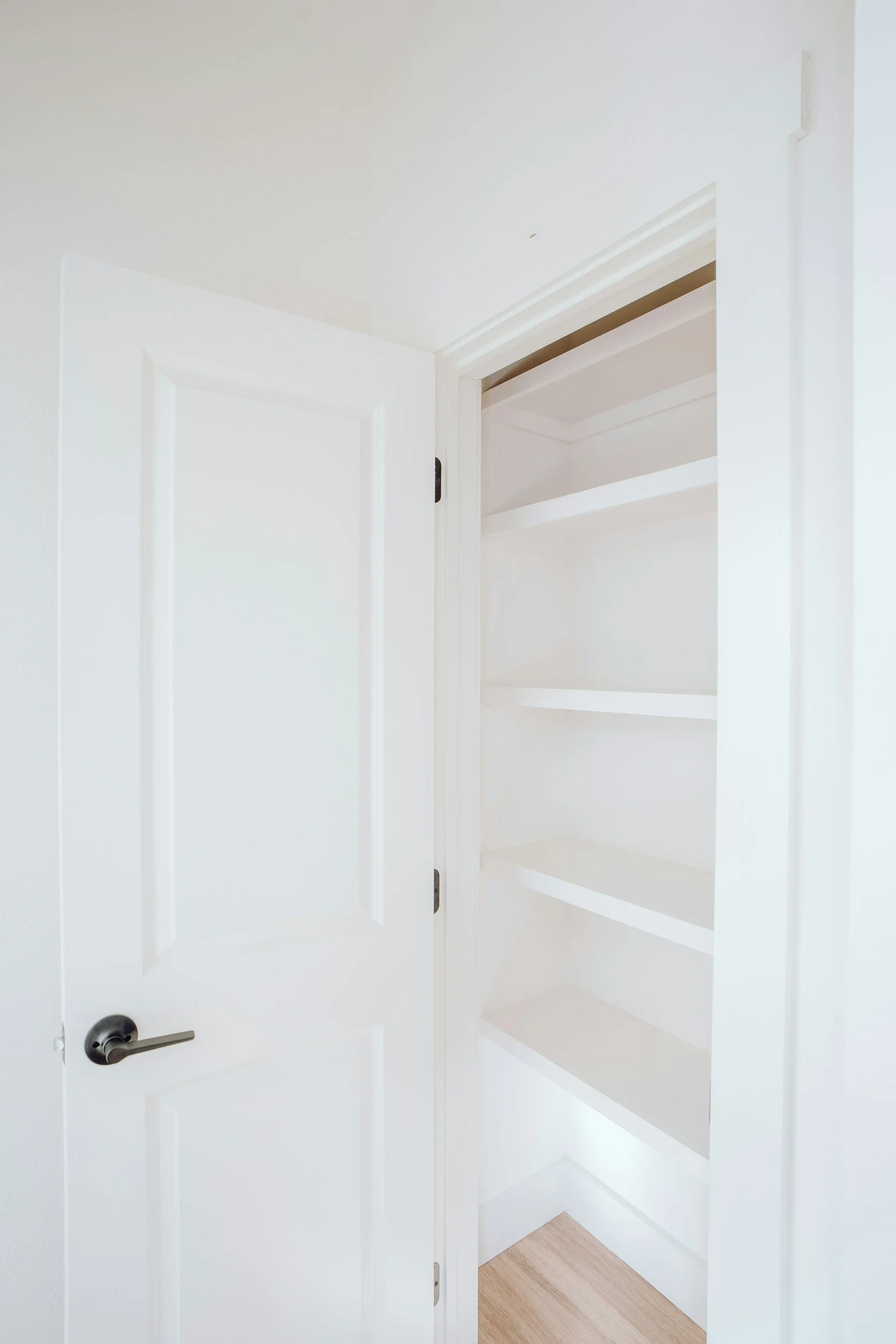Open white closet door revealing empty shelves inside, in a room with light-colored wooden flooring. West Hartford Closet Company WH Closet Co.