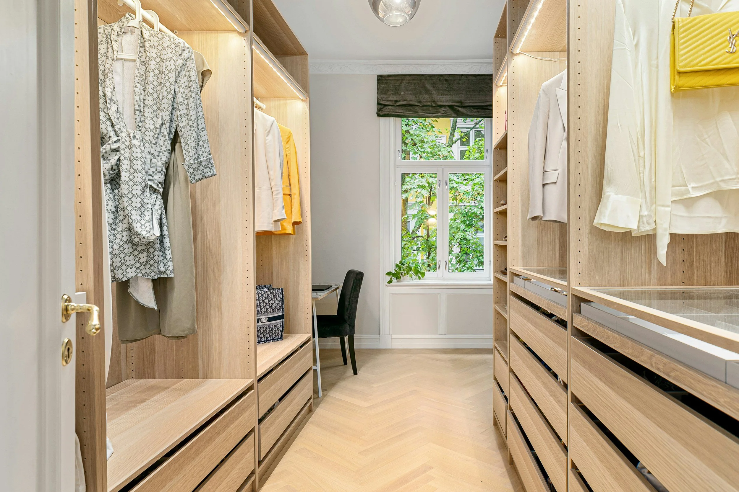A walk-in closet with light wood shelving and drawers, white walls, a large window with a dark valance, and a small desk with a black chair. Clothes and accessories are displayed on the shelving. West Hartford Closet Company WH Closet Co.