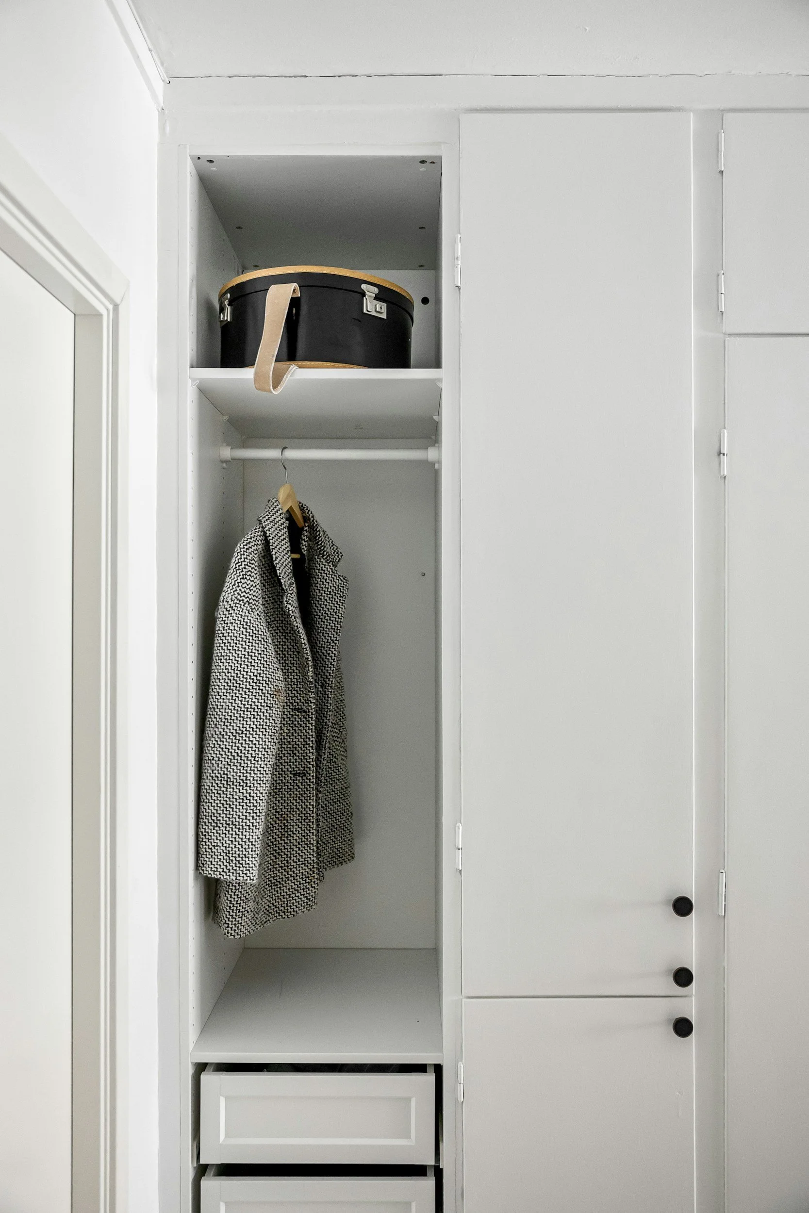 Open white closet with hanging hook holding a checkered coat, a black circular storage box on top shelf, and three black knobs on the right side. West Hartford Closet Company WH Closet Co.