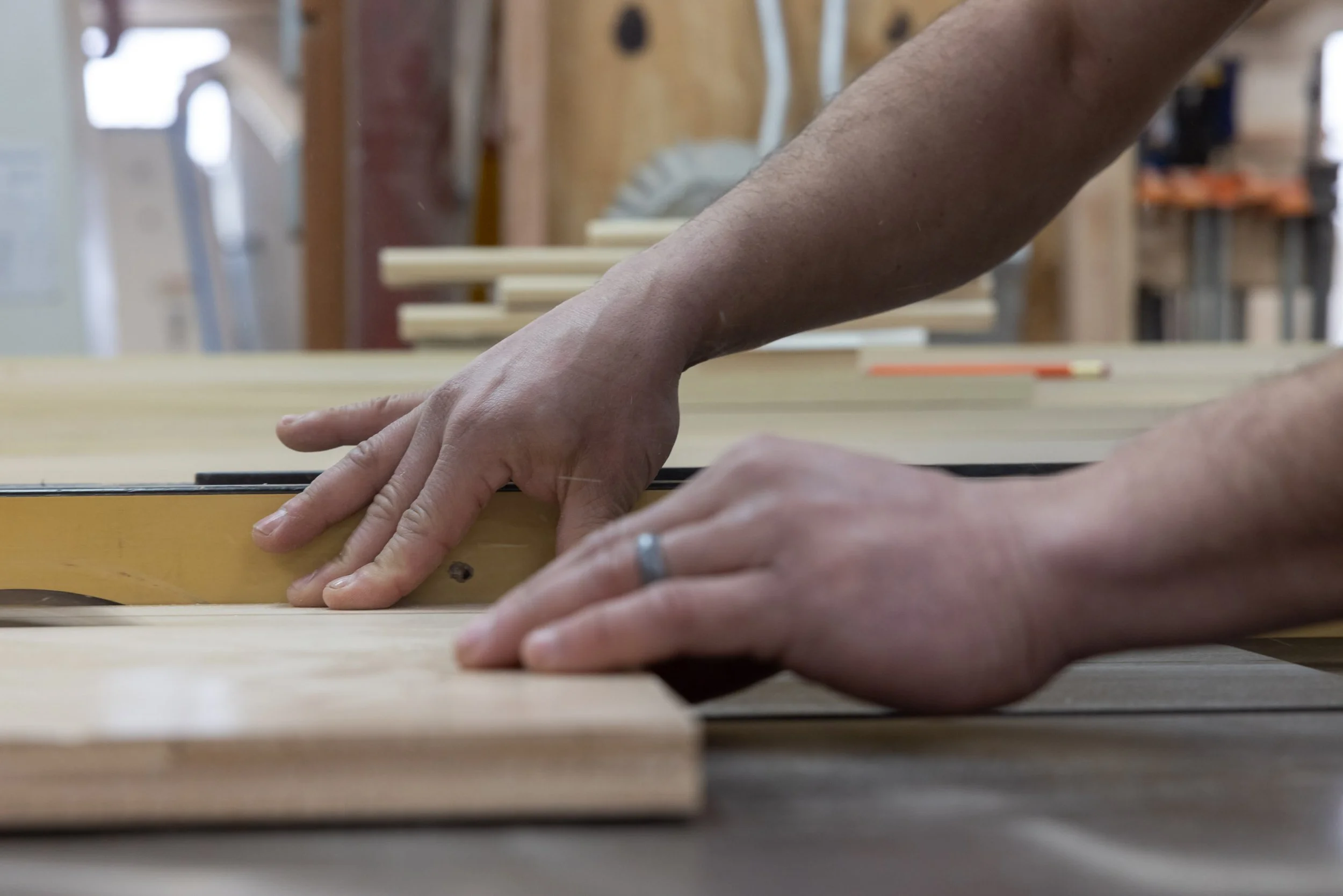 Close-up of a person's hands working with wood, possibly measuring or marking, in a woodworking shop with tools and wooden pieces in the background. West Hartford Closet Company WH Closet Co.
