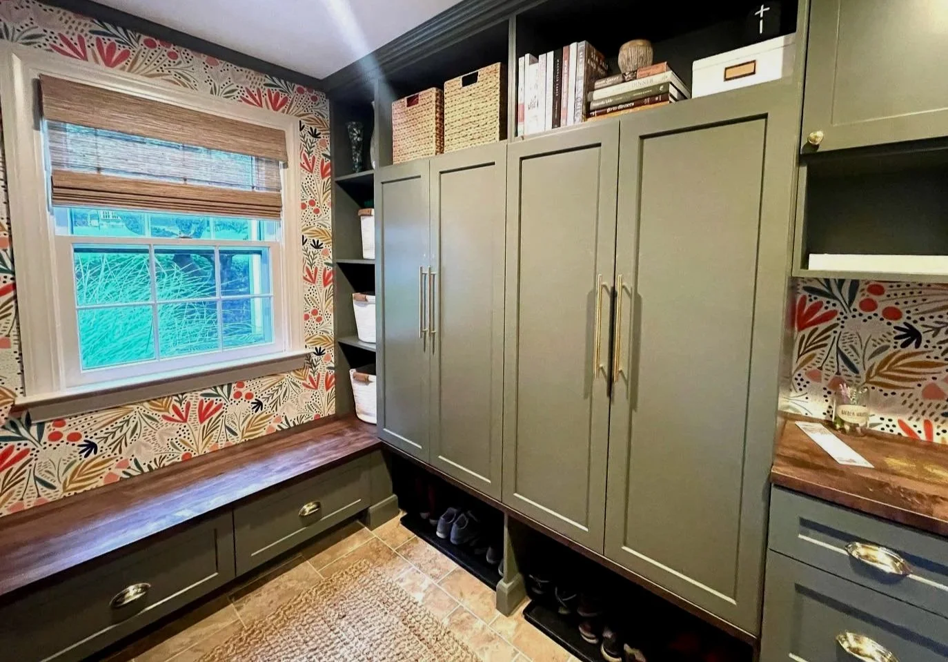 Mudroom with green cabinets, a window with a bamboo shade, a wooden bench, patterned wallpaper, and shelves with baskets and books. West Hartford Closet Company WH Closet Co.