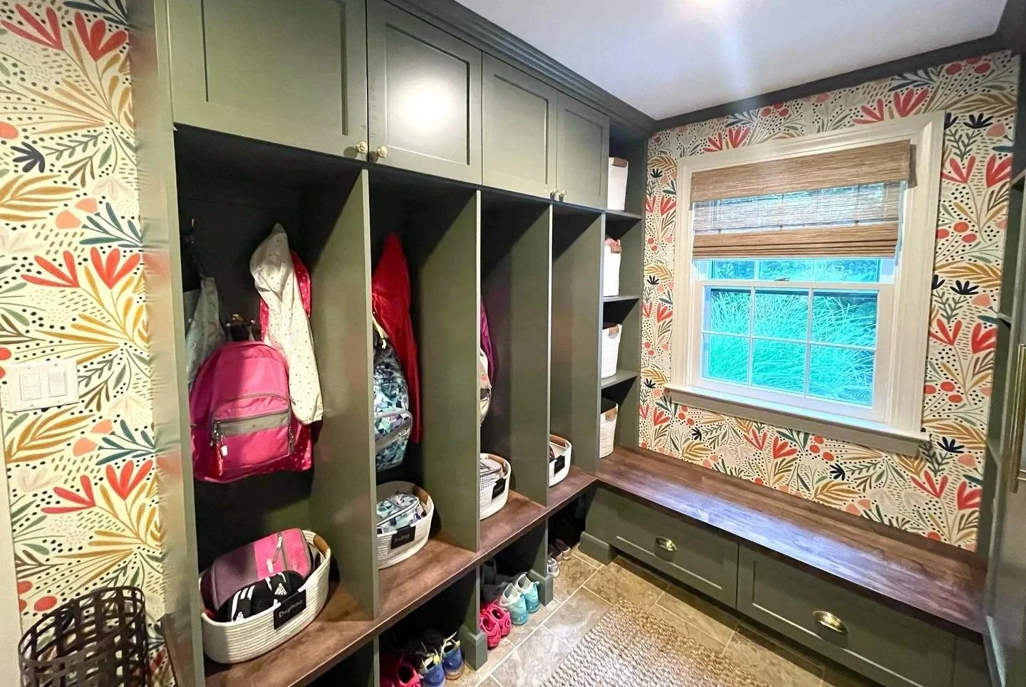 A colorful mudroom with patterned wallpaper, green wall-mounted storage with open cubbies holding backpacks, jackets, and baskets, a window with a woven shade, and a wooden bench with drawers. West Hartford Closet Company WH Closet Co.