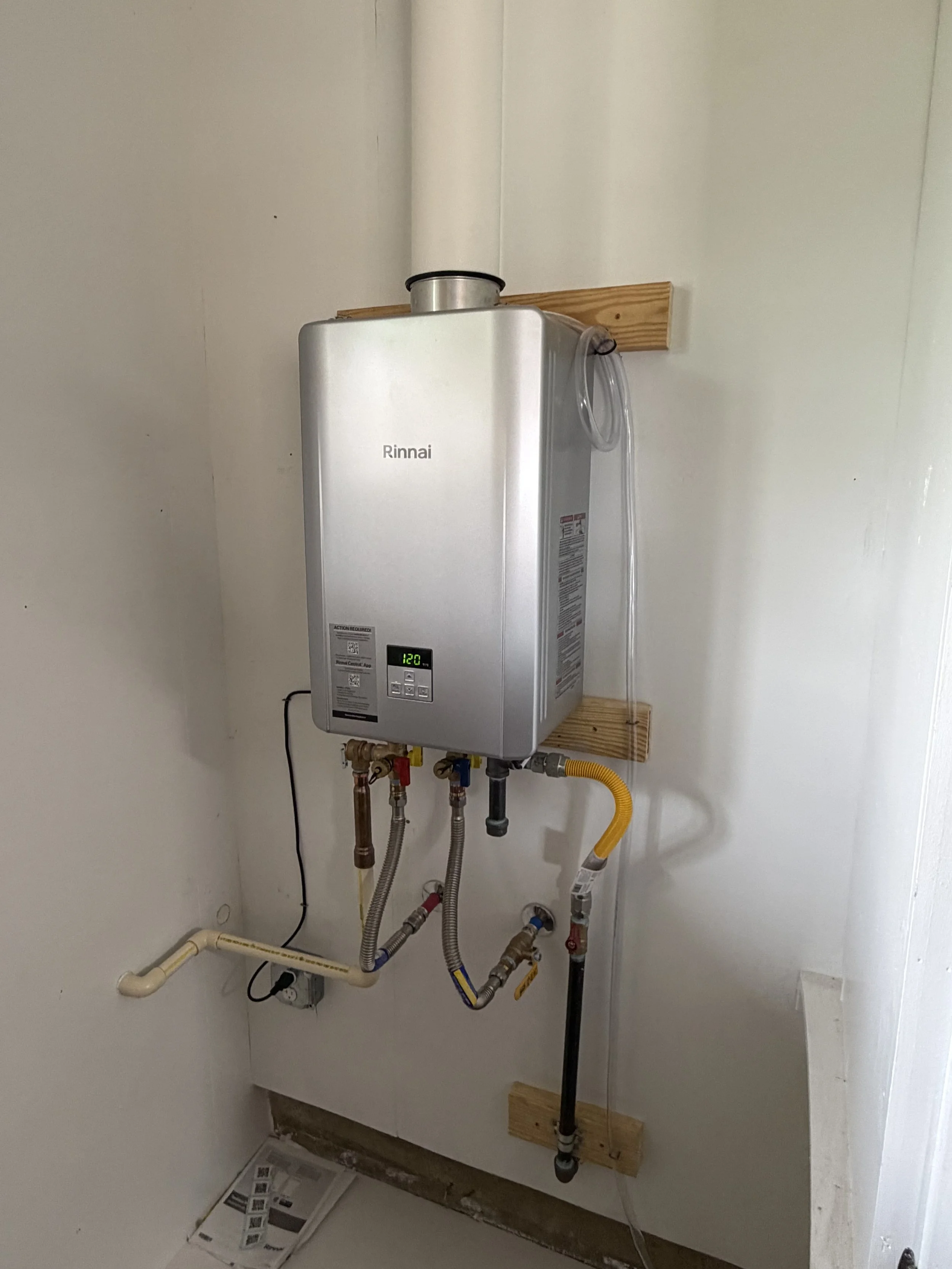 A Rinnai tankless water heater installed on a white wall with plumbing pipes and valves connected at the bottom.