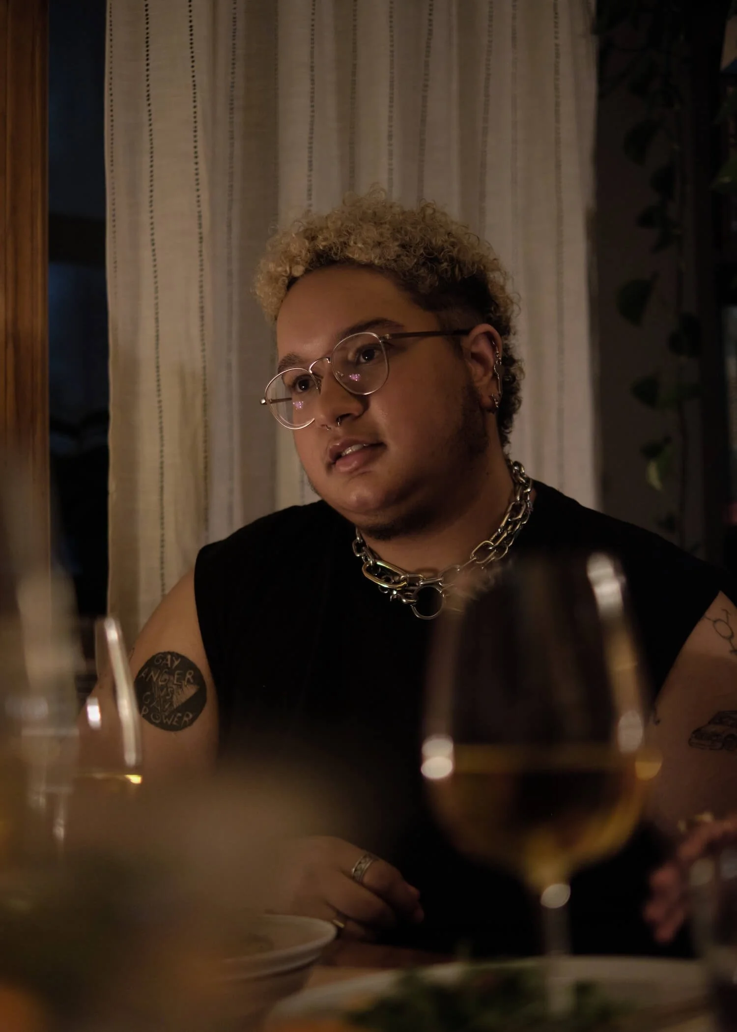 A trans-masculine non-binary person with glasses and blonde curly hair sitting at a dining table with wine glasses and a wine bottle, indoors near curtains.