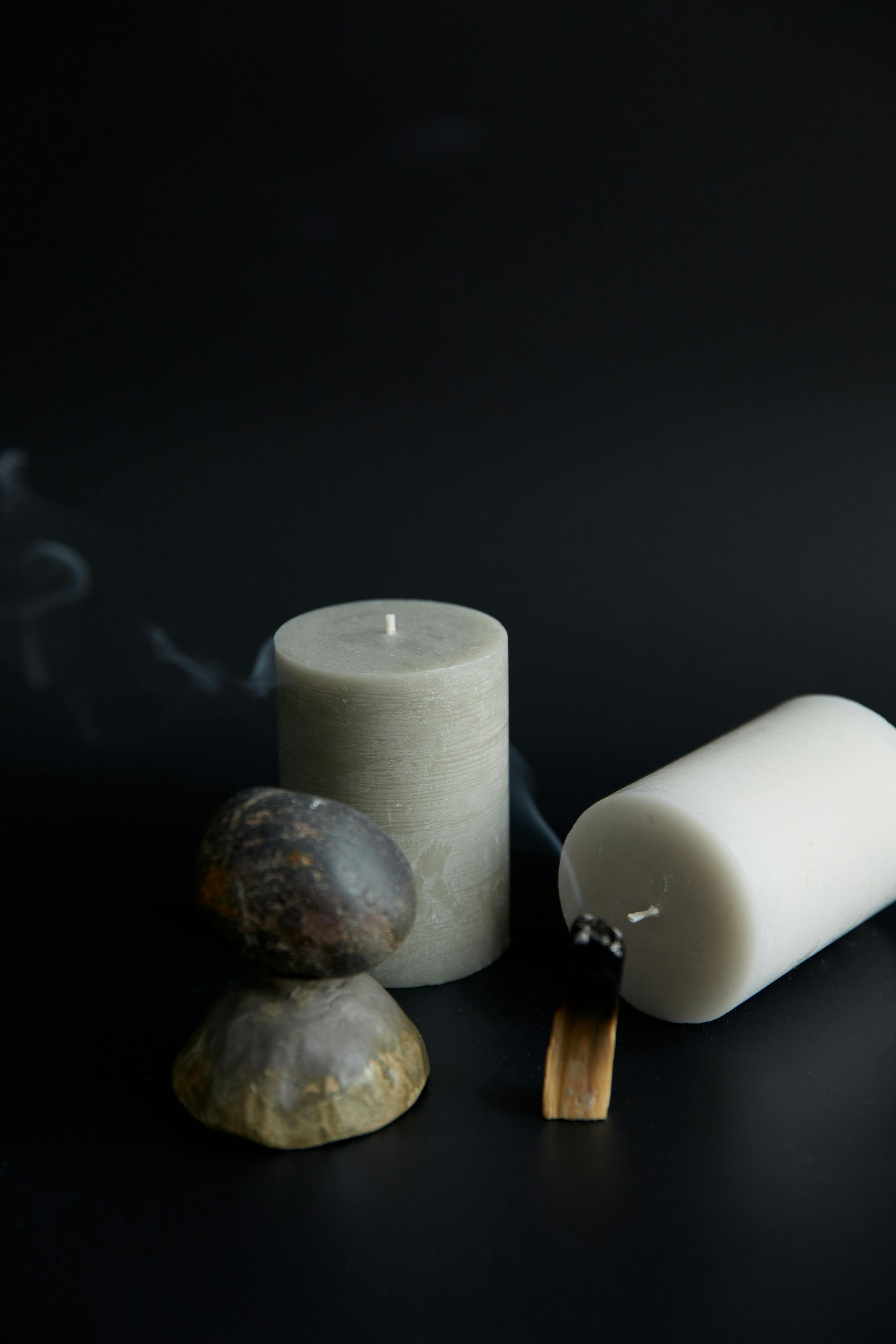 A still life arrangement of two white candles, one tall and one short, with two stacked stones and a burnt matchstick on a dark background.