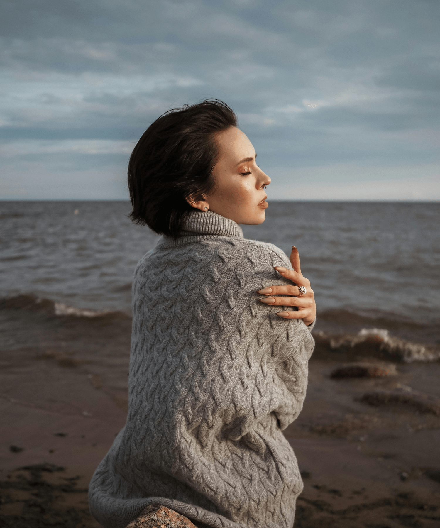 A woman with short dark hair stands on the beach near the water, wearing a gray knitted sweater, with her eyes closed and arms crossed over her chest.