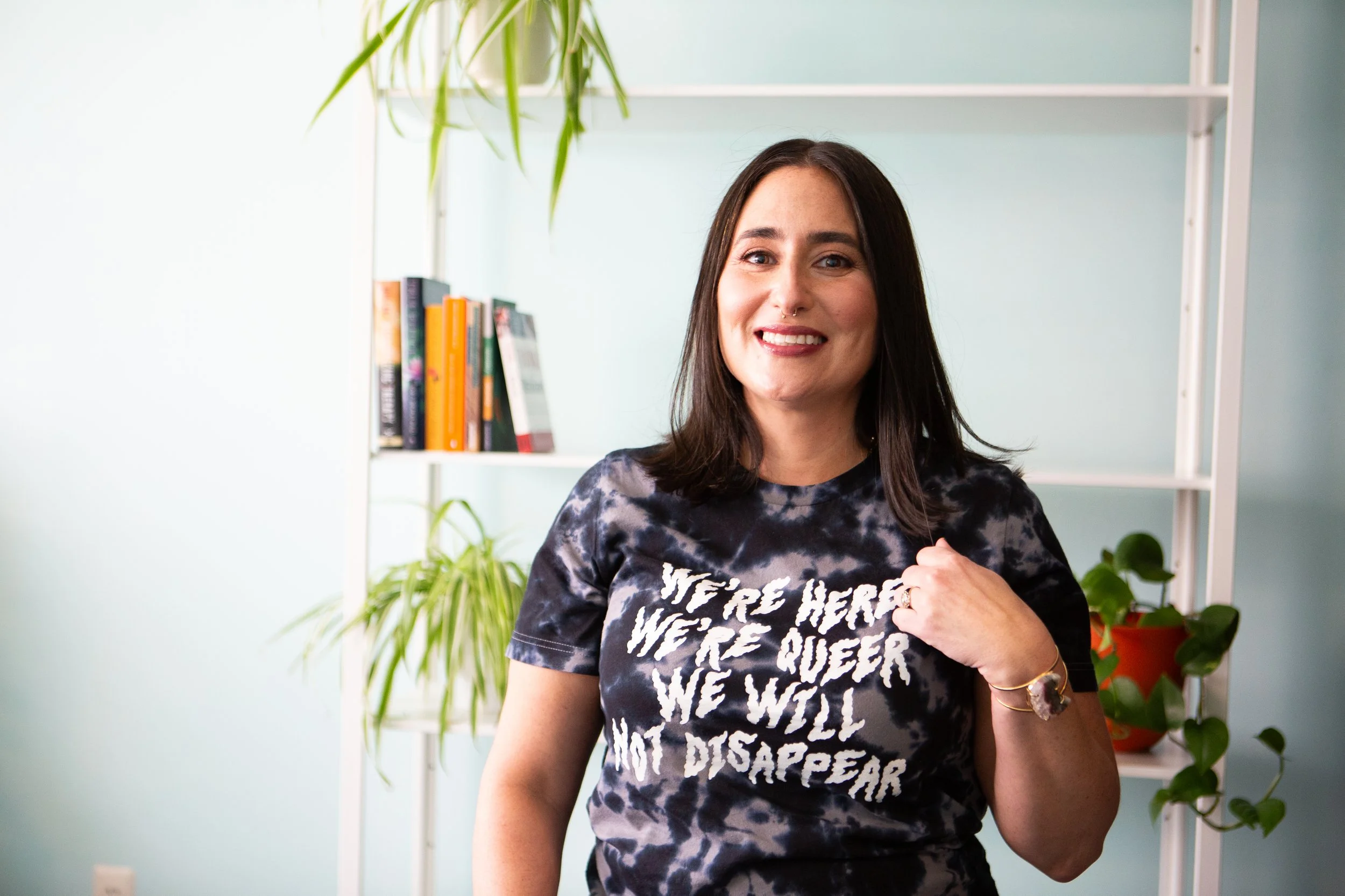A woman with shoulder-length dark hair is smiling and wearing a black and white tie-dye t-shirt with the message 'WE'RE HERE, WE'RE QUEER, WE WILL NOT DISAPPEAR.' She is standing in a room with white shelves and green plants in the background.